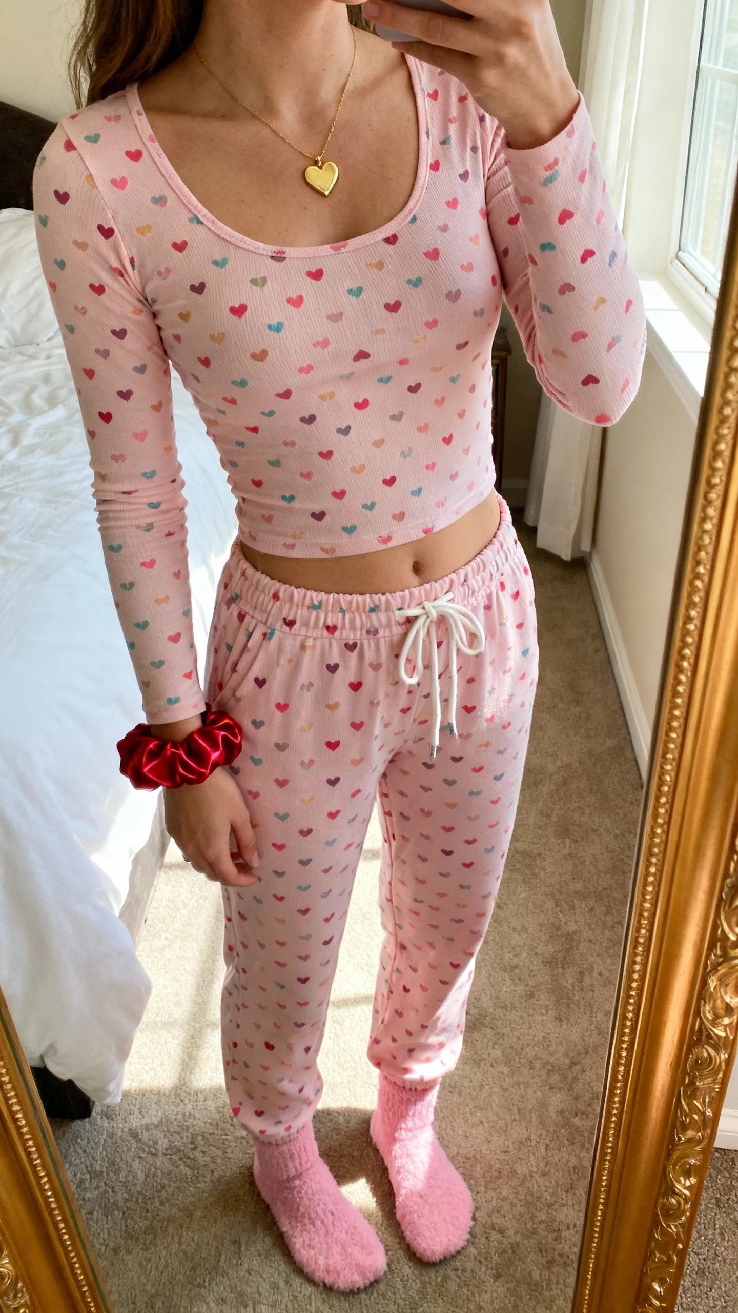 iPhone mirror selfie of a woman wearing matching candy-heart print pajama set with a chic twist: fitted long-sleeve top with tiny candy-heart pattern, high-waist drawstring pants, soft pink fuzzy socks, delicate gold heart pendant, and a red satin scrunchie on wrist; face not visible, cropped at neck, bedroom mirror with morning window light, iPhone photo quality.