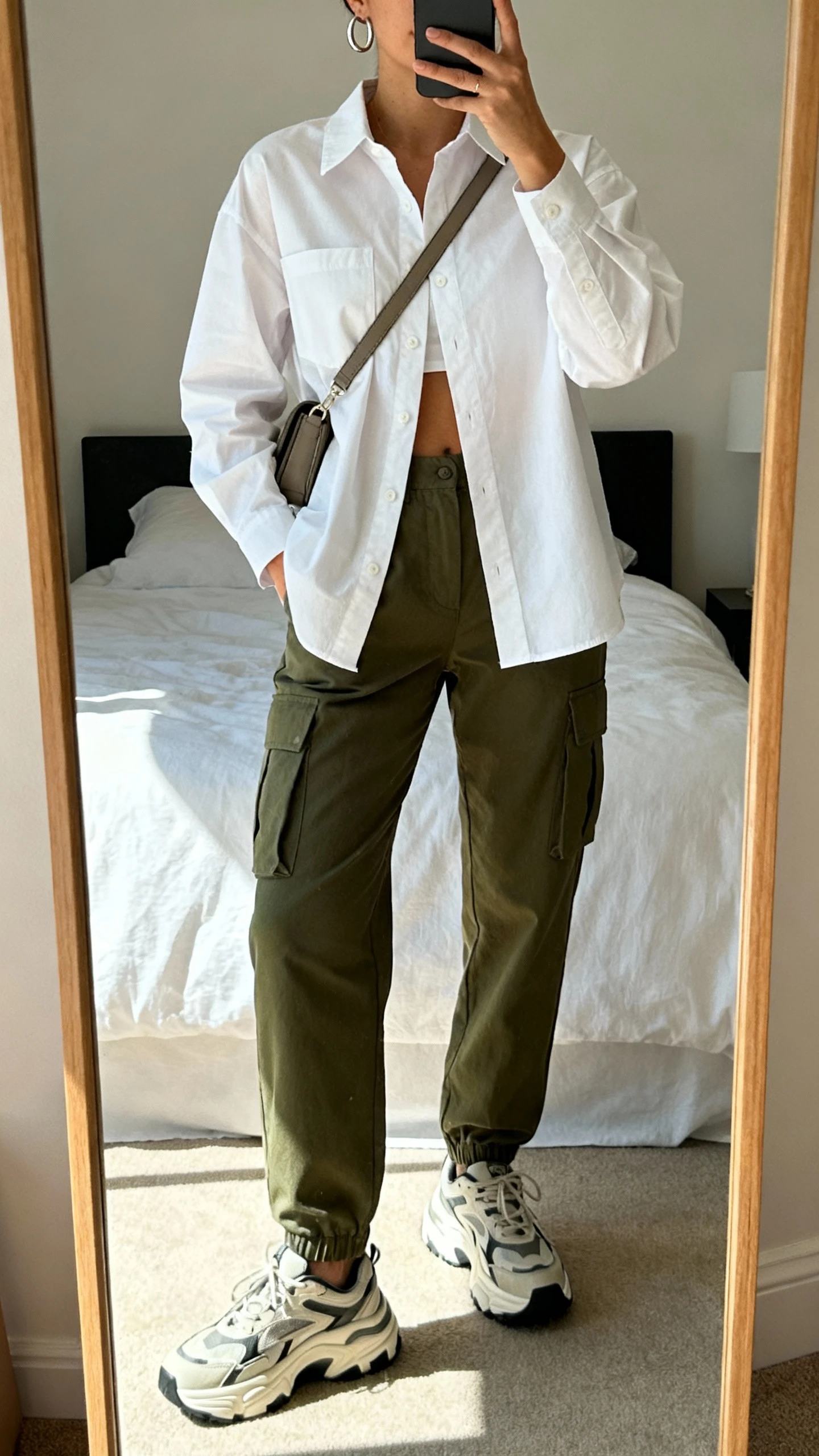 iPhone mirror selfie of a woman wearing olive cargo trousers, a crisp white button-up shirt half-tucked, chunky sneakers, a crossbody bag, and minimalist hoop earrings, face not visible, cropped at shoulders, bedroom mirror with soft afternoon light, iPhone photo quality.