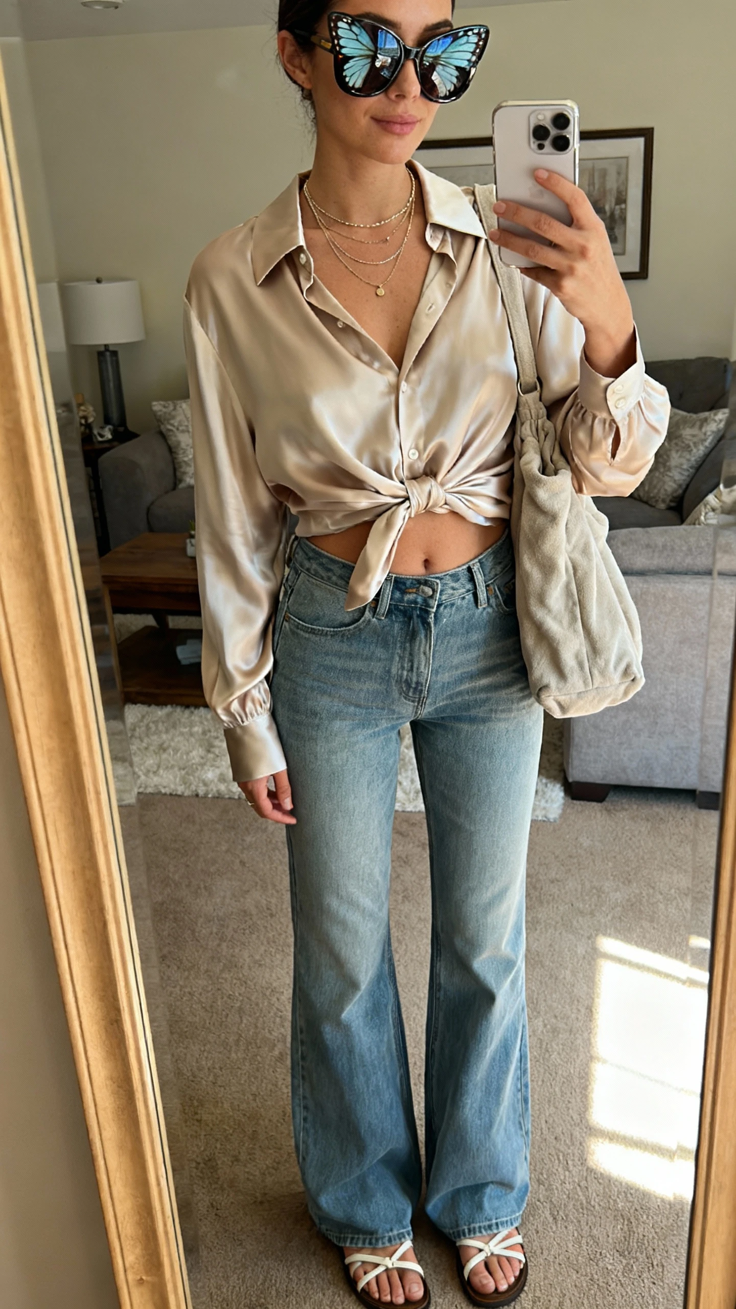 iPhone mirror selfie of a woman wearing oversized butterfly sunglasses, a drapey satin blouse half-tucked into relaxed flare jeans, strappy sandals, layered delicate necklaces, and a soft slouchy shoulder bag, face not visible, cropped at neck, living room mirror, warm indoor lighting, iPhone photo quality.