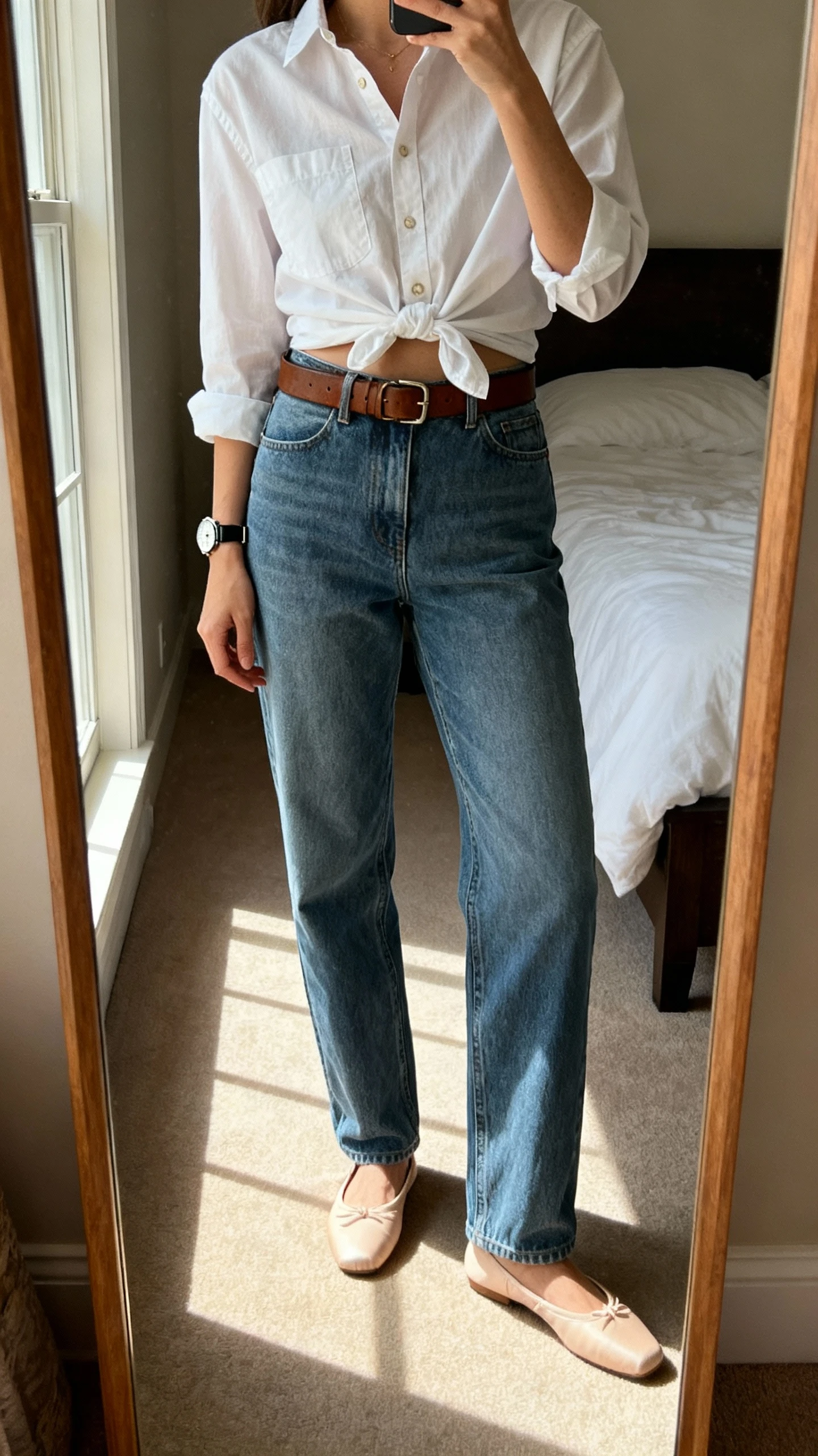 iPhone mirror selfie of a woman wearing relaxed straight-leg jeans, crisp button-down shirt half-tucked, leather belt, ballet flats, and a classic watch, face not visible, cropped at shoulders, bedroom mirror with morning window light, iPhone photo quality.