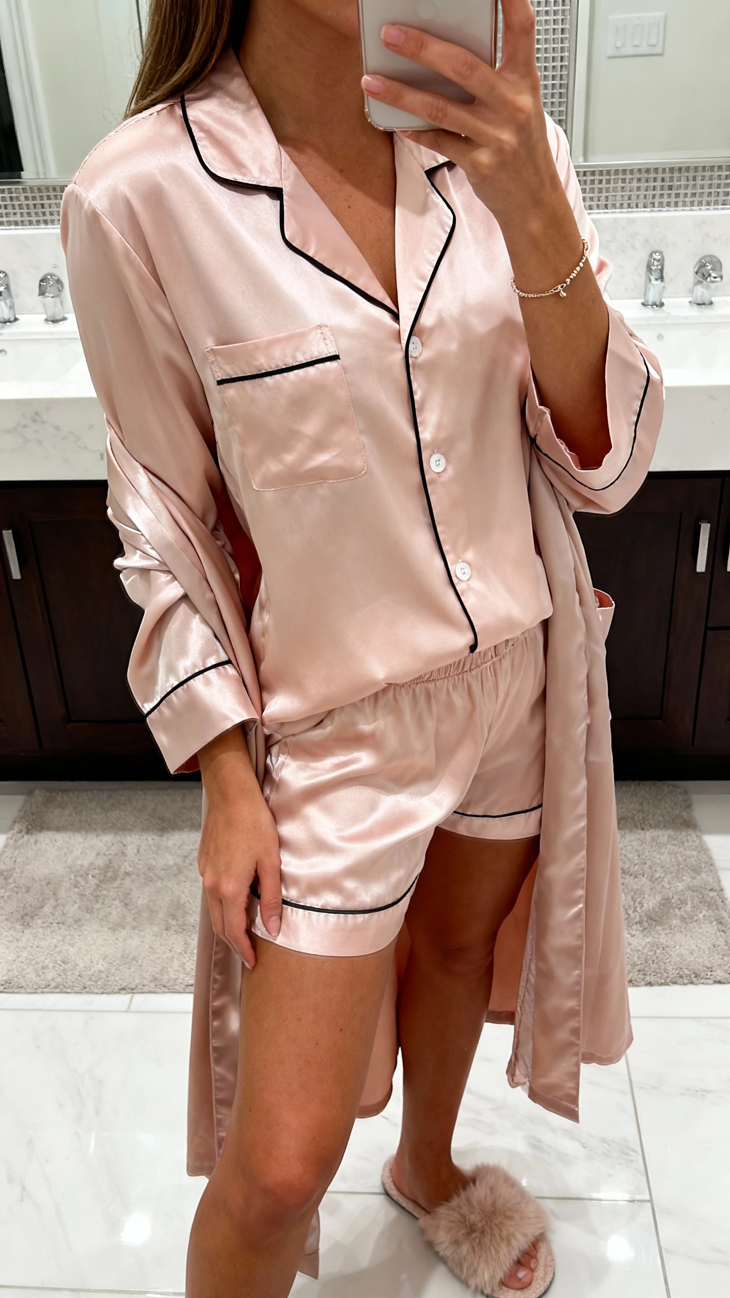 iPhone mirror selfie of a woman wearing satin sweethearts set: blush satin button-up pajama shirt with black piping, matching satin shorts, silky robe draped open, delicate bracelet, fluffy slippers; face not visible, side angle, bathroom mirror with bright indoor lighting, iPhone photo quality.