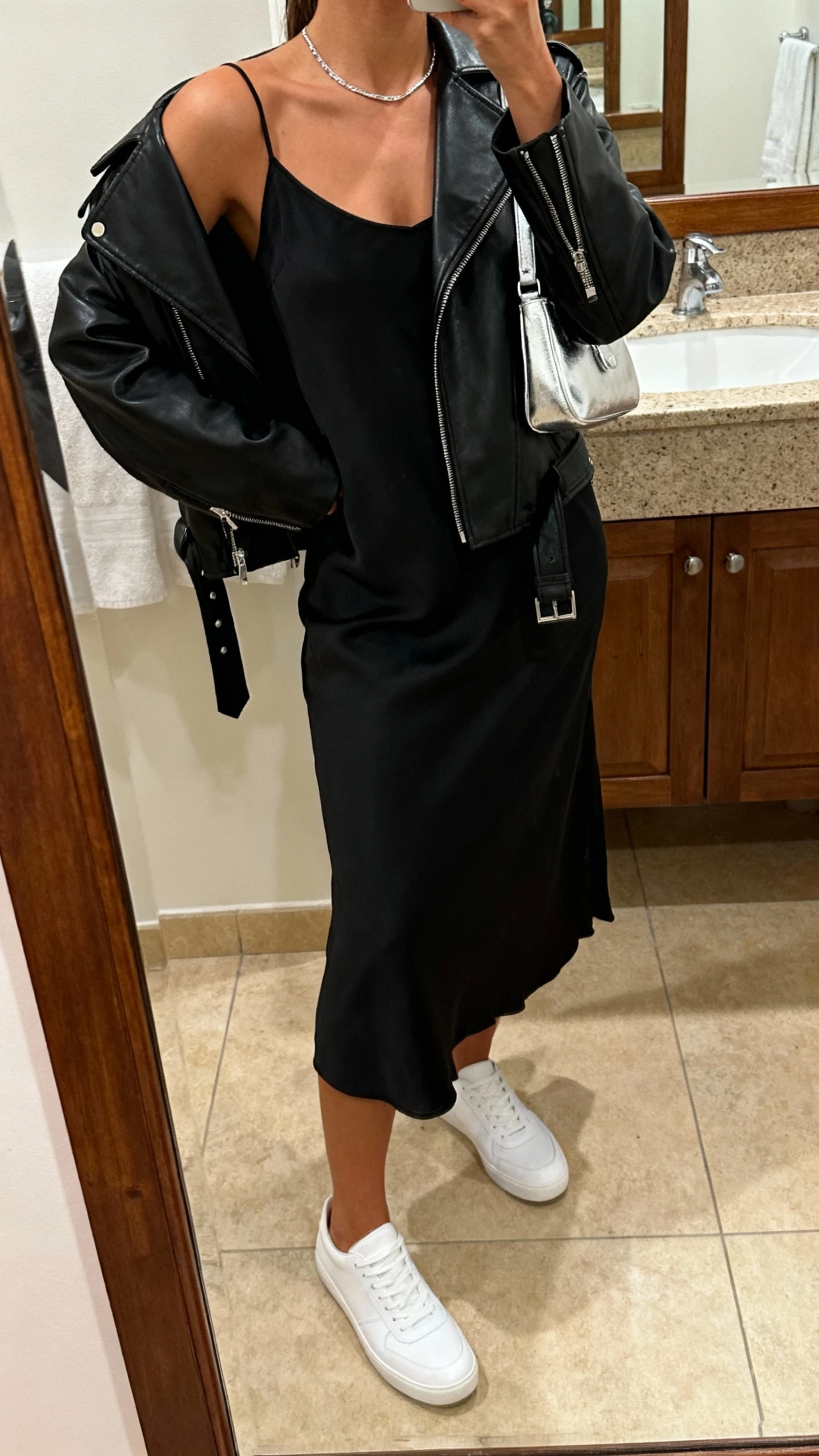iPhone mirror selfie of a woman wearing sleek white minimalist sneakers, a black slip dress with a black leather moto jacket draped over shoulders, a small silver shoulder bag, and delicate silver necklace, face not visible, cropped at neck, bathroom mirror under warm indoor lighting, iPhone photo quality.