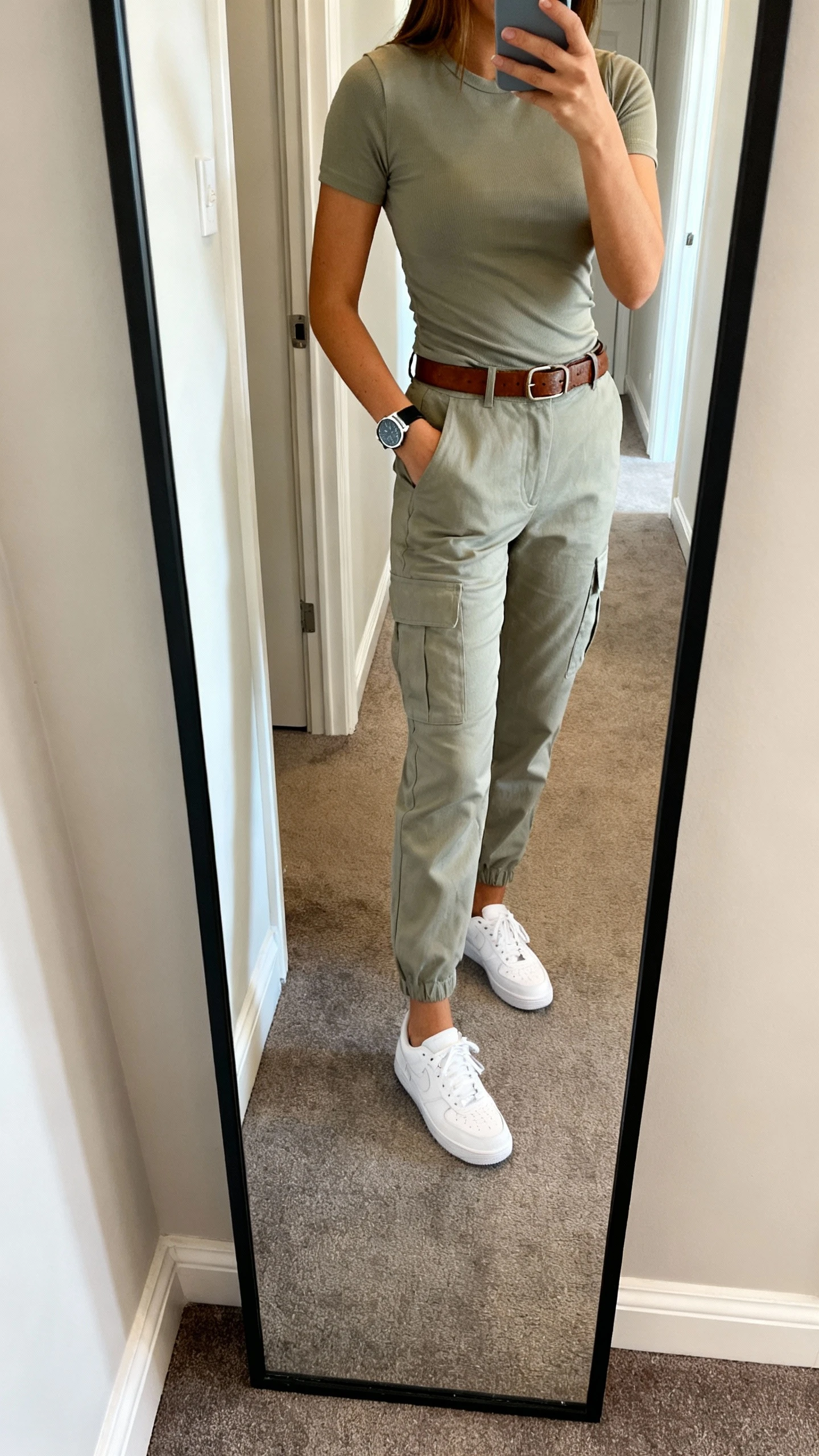 iPhone mirror selfie of a woman wearing soft cargo pants with an elevated fitted tee, leather belt, clean white trainers, and a sleek watch, face not visible, cropped at shoulders, full-length hallway mirror with natural daylight, iPhone photo quality.