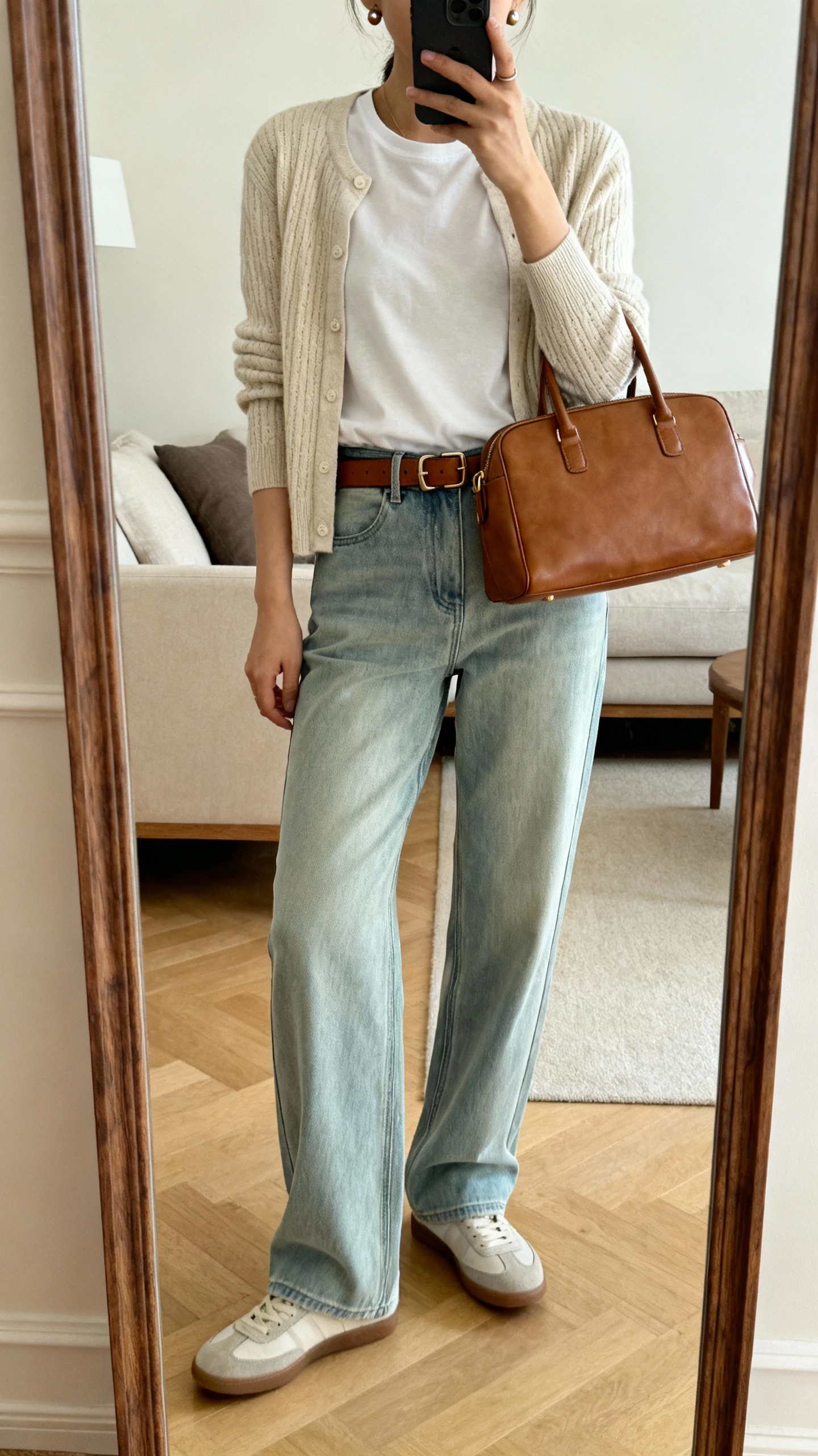 iPhone mirror selfie of a woman wearing soft relaxed jeans, a fine-knit cardigan over a simple tee, quiet-luxury leather belt, minimalist sneakers, small stud earrings, and a structured leather tote; face not visible, cropped at shoulders, full-length mirror in living room, warm indoor lighting, iPhone photo quality.