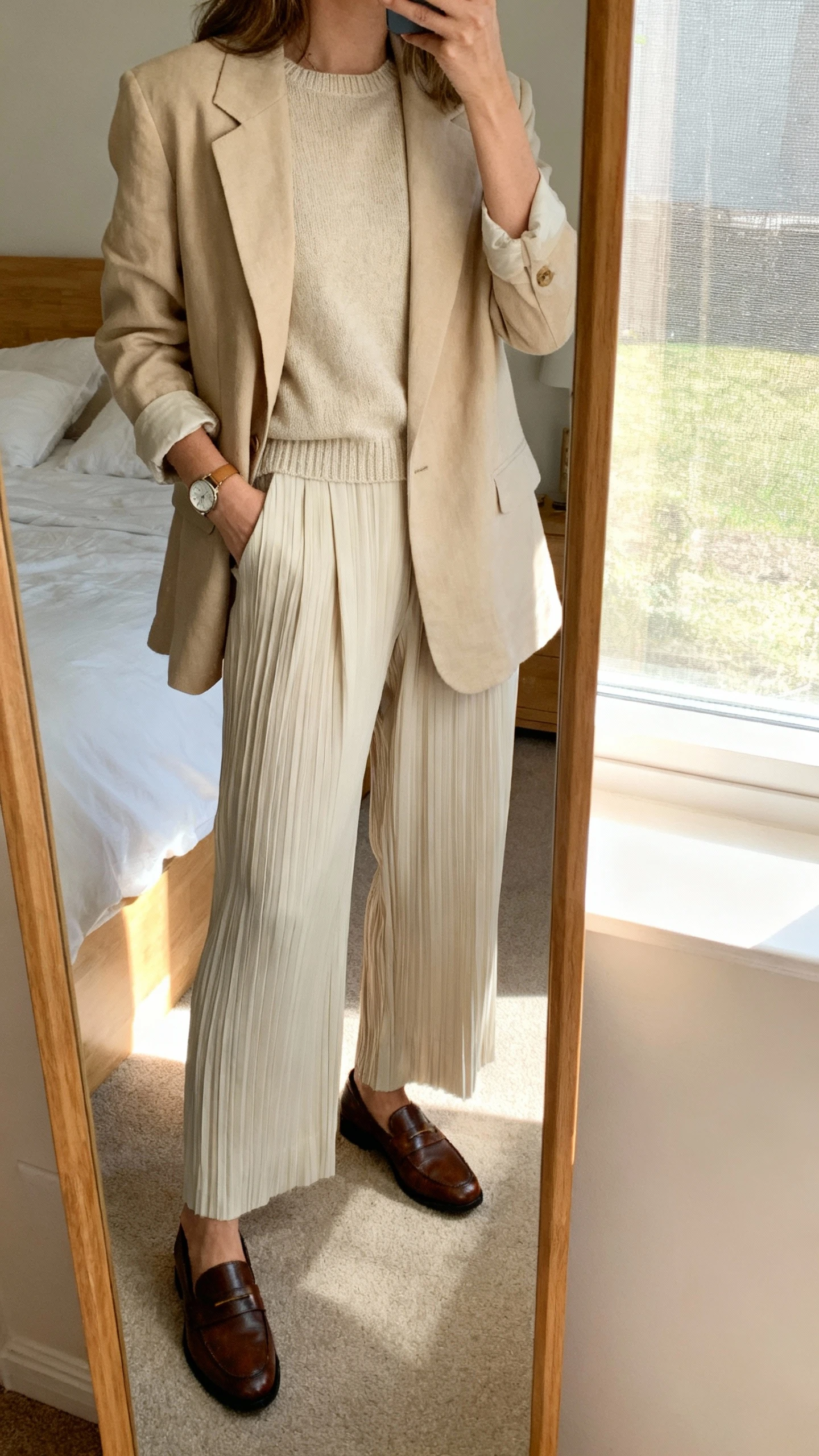 iPhone mirror selfie of a woman wearing soft tailoring: relaxed beige blazer over a fine-knit crewneck, pleated wide-leg trousers, classic leather loafers, delicate watch, face not visible, cropped at shoulders, bedroom mirror with window light, iPhone photo quality.