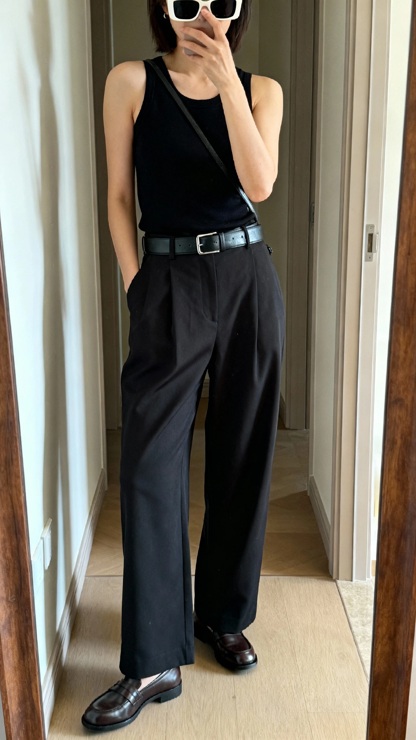 iPhone mirror selfie of a woman wearing square sunglasses, a minimalist black tank, high-waisted wide-leg trousers, sleek leather belt, chunky slip-on loafers, and a slim crossbody, face not visible, cropped at shoulders, full-length mirror in hallway, indoor daylight, iPhone photo quality.