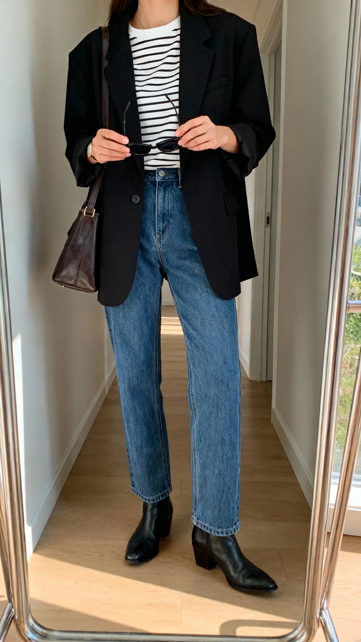 iPhone mirror selfie of a woman wearing straight-leg blue jeans, a black oversized blazer over a striped tee, ankle boots, and a leather shoulder bag with sunglasses in hand, face not visible, cropped at shoulders, full-length hallway mirror, natural afternoon light, iPhone photo quality.