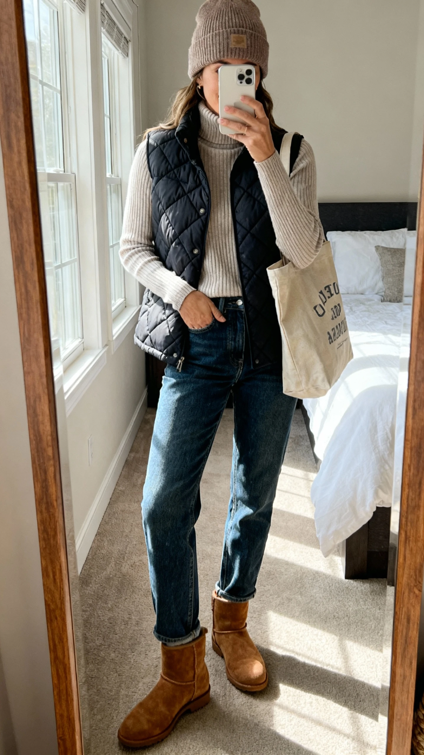 iPhone mirror selfie of a woman wearing suede desert boots, relaxed straight-leg jeans, cozy ribbed turtleneck, quilted vest, beanie, and a canvas tote, face not visible, cropped at shoulders, bedroom mirror with soft afternoon window light, iPhone photo quality.