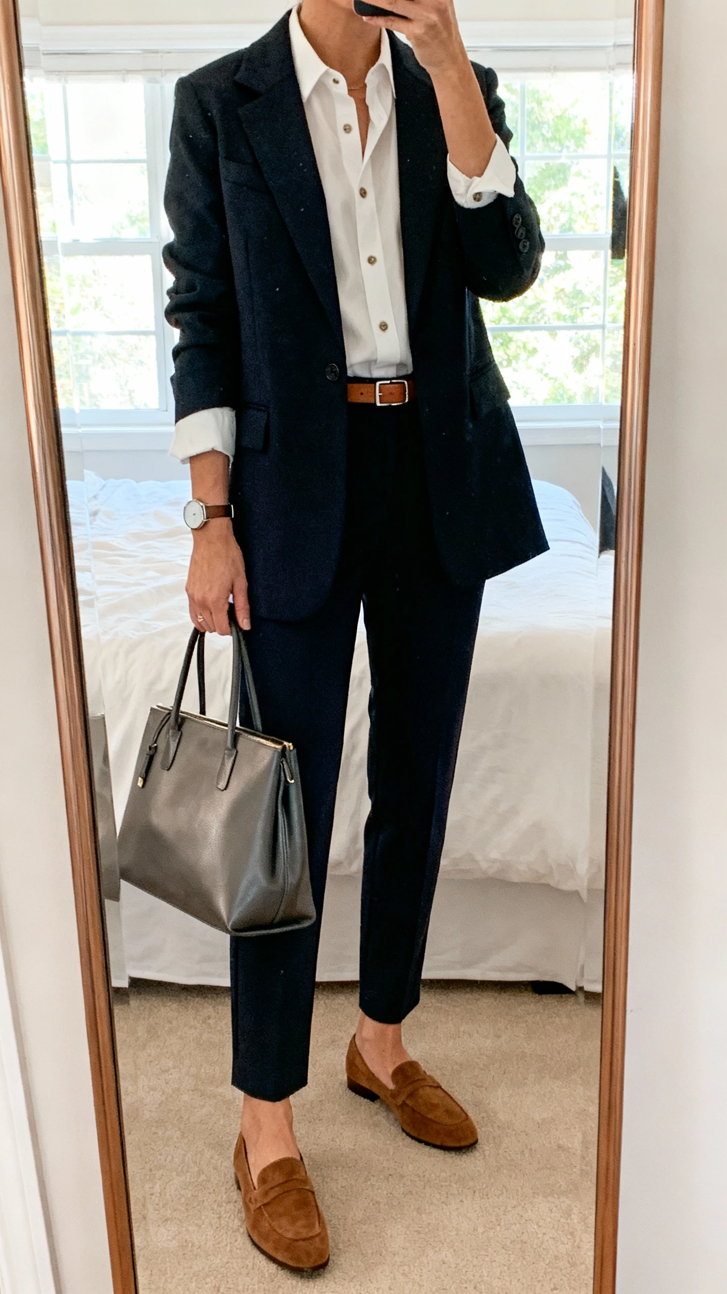 iPhone mirror selfie of a woman wearing suede loafers, tailored ankle trousers, crisp button-up shirt under a structured blazer, slim leather belt, minimalist watch, and a sleek tote, face not visible, cropped at neck, bedroom mirror with natural window light, iPhone photo quality.