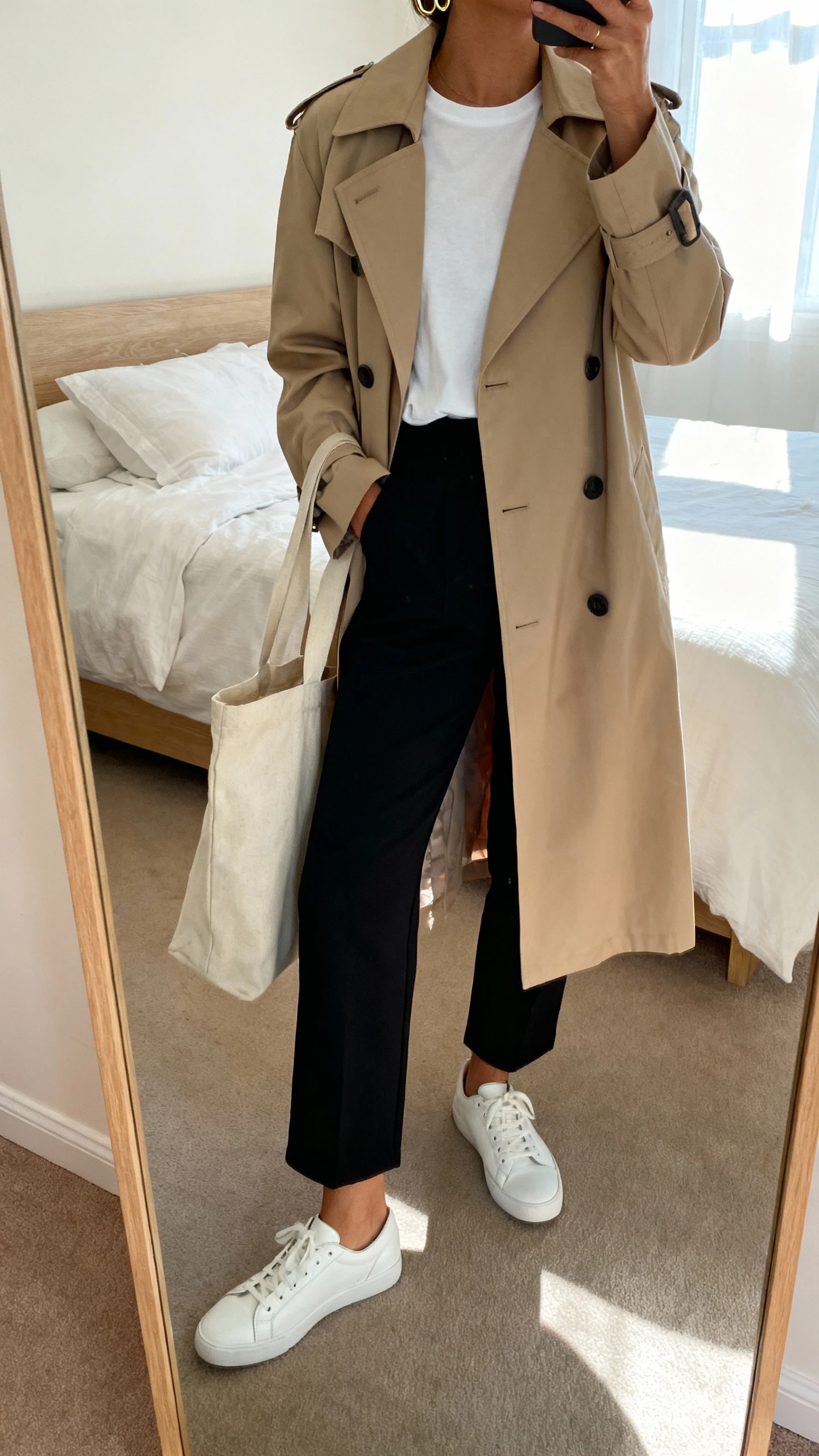 iPhone mirror selfie of a woman wearing white leather low-top sneakers, a tailored beige trench coat over a white tee, black ankle-length trousers, a canvas tote, and minimal gold hoops, face not visible, cropped at neck, bedroom mirror with morning window light, iPhone photo quality.