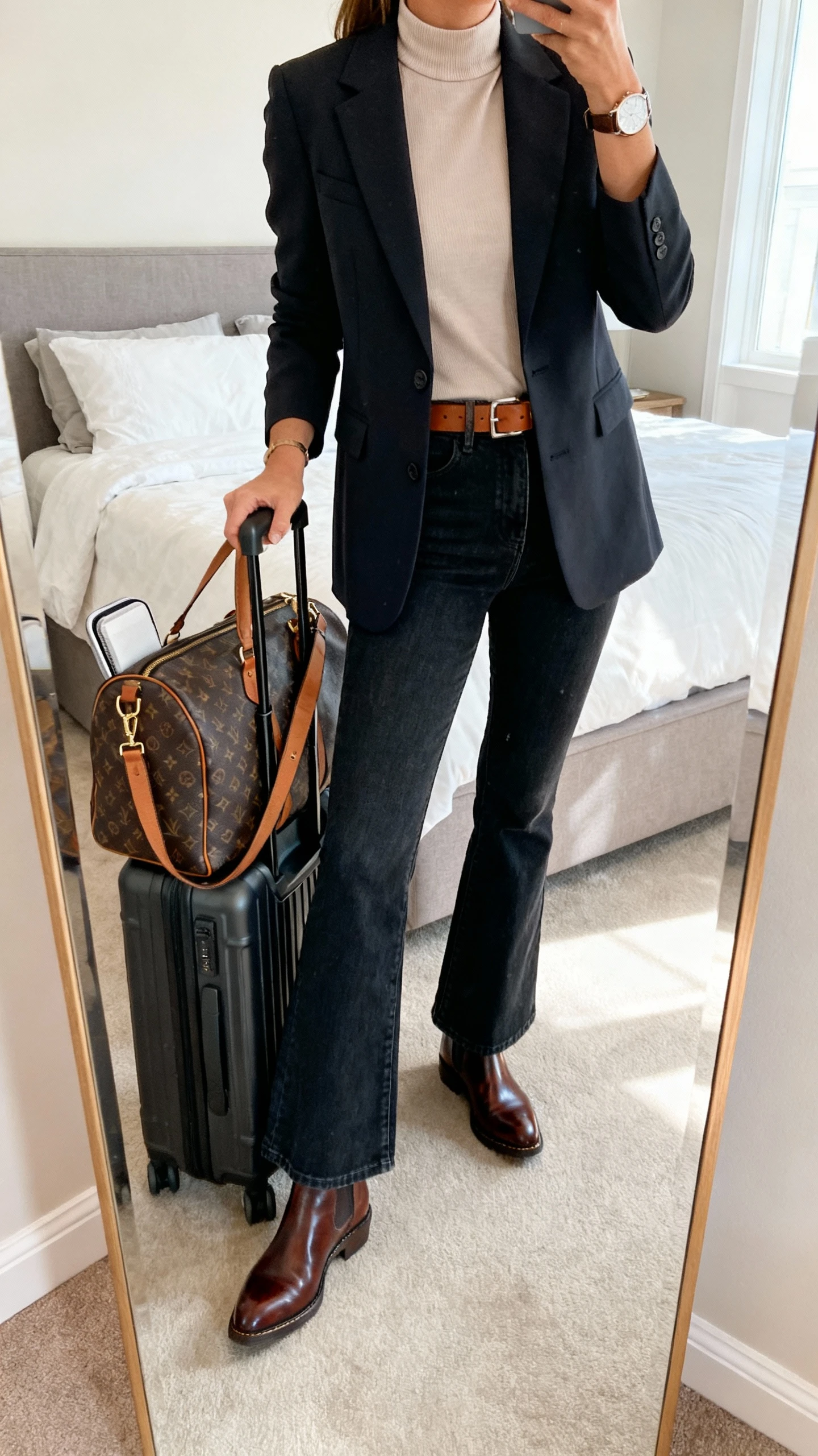 iPhone mirror selfie of a woman with a medium weekender carryall with trolley sleeve and crossbody strap, a breathable mock-neck top, high-stretch flare jeans, polished Chelsea boots, a tailored blazer, and a minimal leather belt and watch, face not visible, cropped at neck, bedroom mirror, soft indoor lighting, iPhone photo quality.