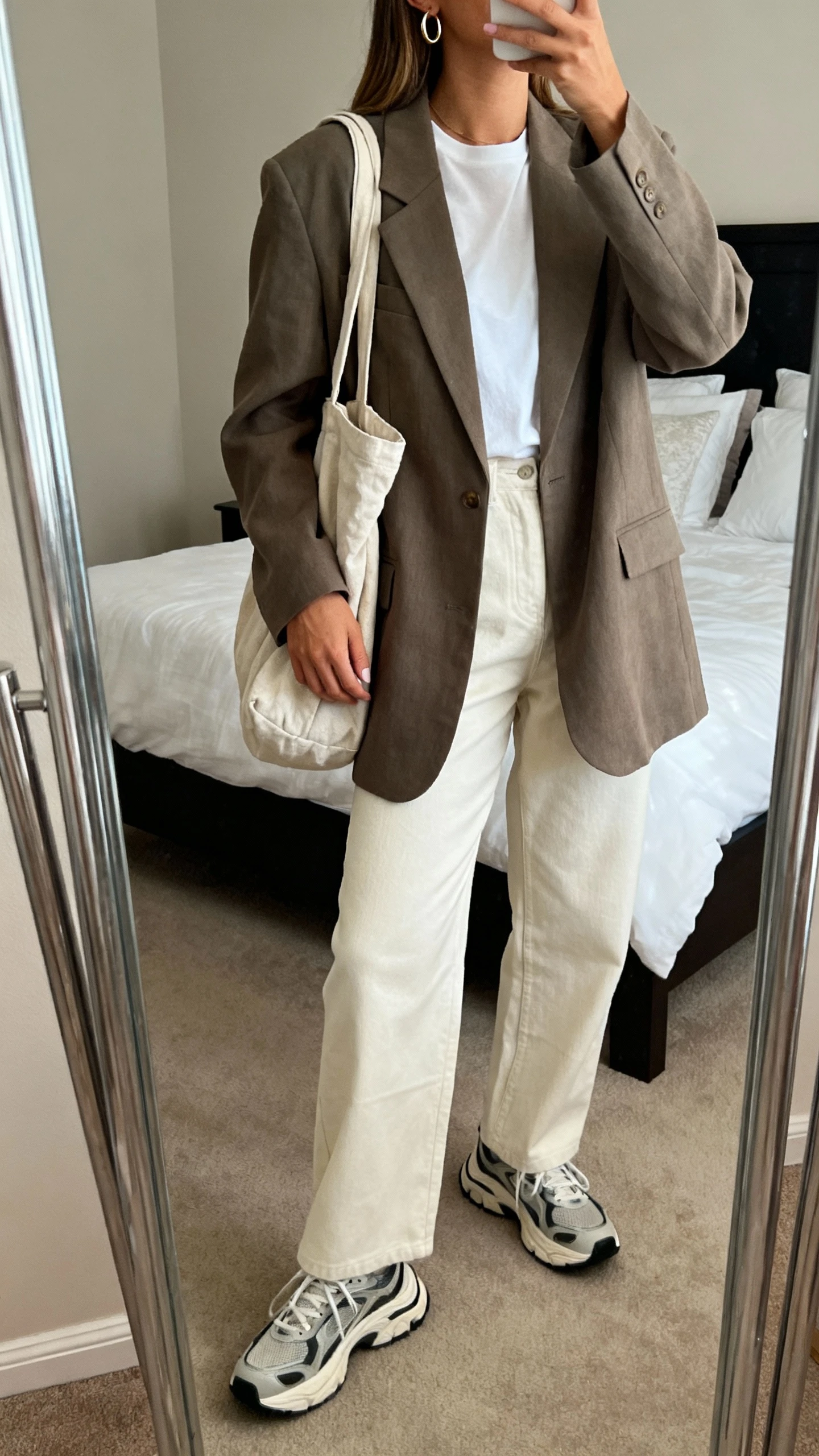 iPhone mirror selfie of a woman with a slouchy tote, oversized structured blazer over a relaxed white tee, wide-leg trousers, chunky retro sneakers, and thin hoop earrings; face not visible, shoulder-crop, bedroom mirror, soft indoor light, iPhone photo quality.