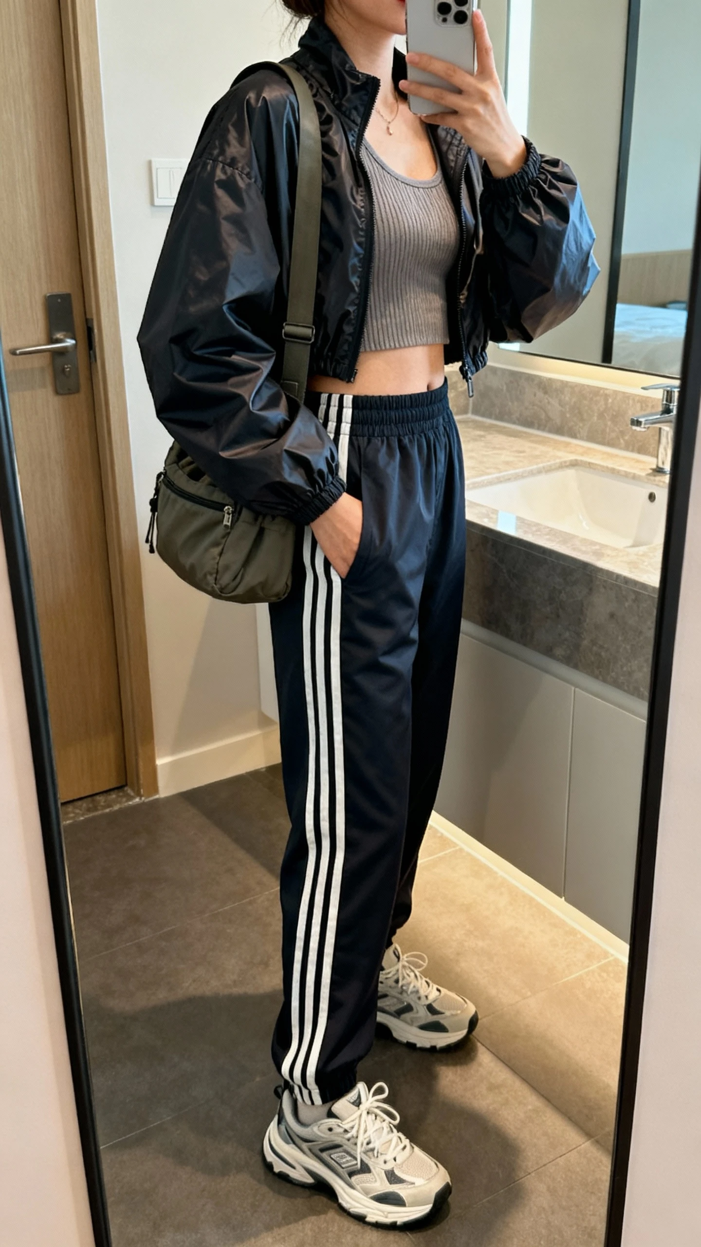 iPhone mirror selfie side angle of a woman in a boxy sporty-cool look: cropped nylon windbreaker, ribbed tank, high-waist track pants with stripes, chunky sneakers, and a nylon shoulder bag; face not visible, bathroom mirror, indoor lighting, iPhone photo quality.