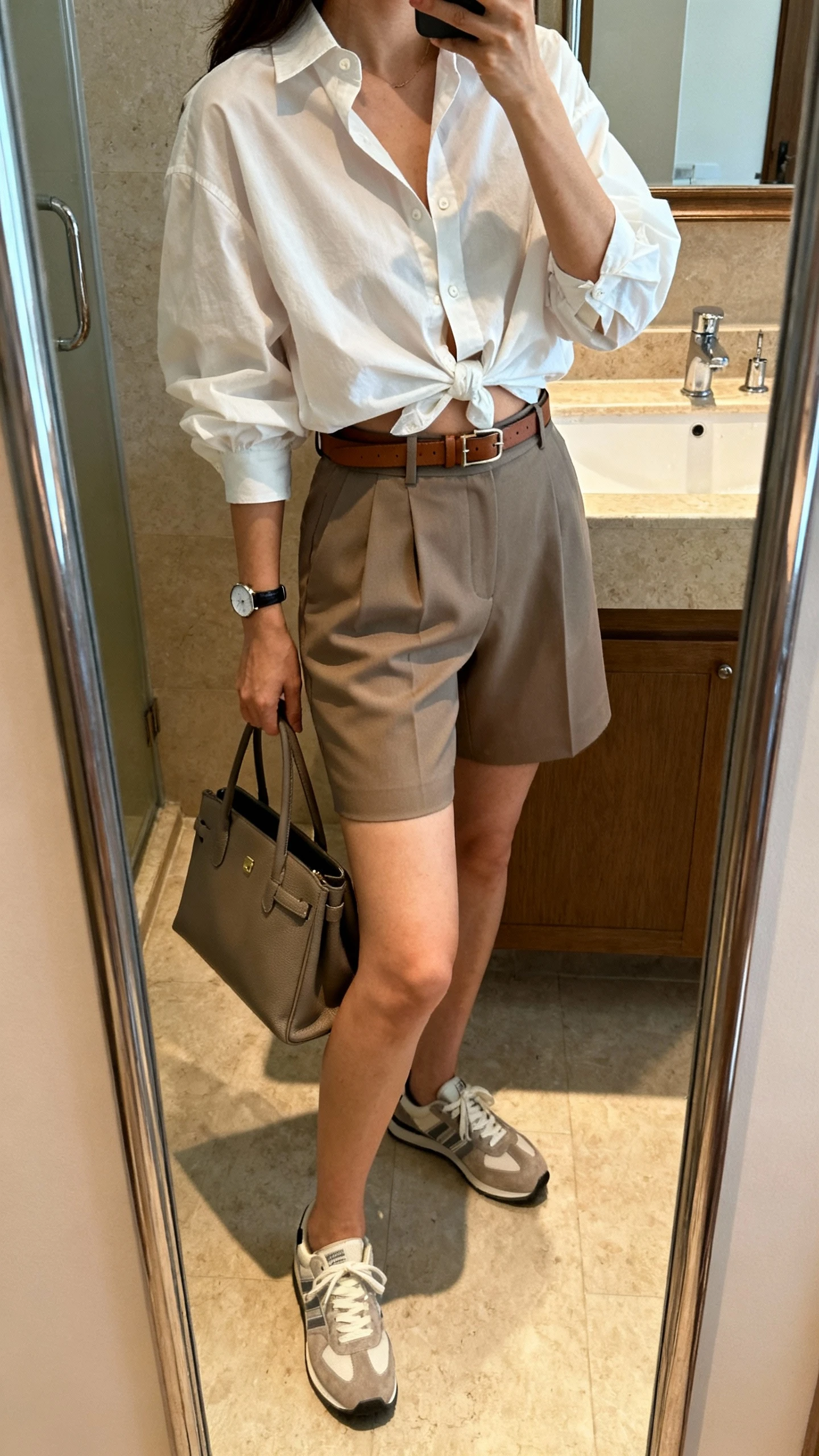 iPhone mirror selfie side angle of a woman wearing tailored high-waist shorts, crisp oversized button-up shirt half-tucked, slim leather belt, comfy low-profile sneakers, structured tote, simple watch, face not visible, bathroom full-length mirror, natural indoor lighting, iPhone photo quality