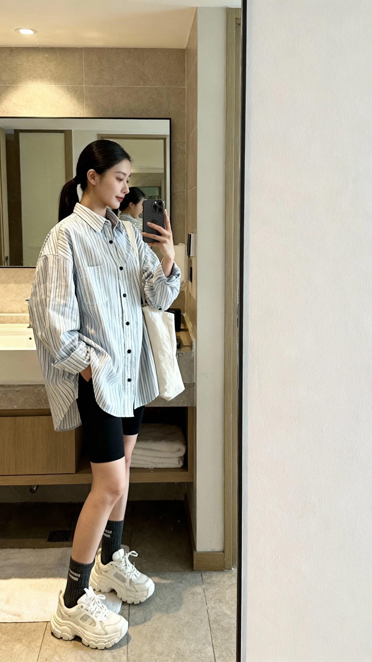 iPhone mirror selfie side angle of a woman wearing an oversized striped button-down shirt with black bike shorts, crew socks with chunky white sneakers, sleek ponytail, simple tote bag, face not visible, bathroom full-length mirror, morning light, iPhone photo quality.