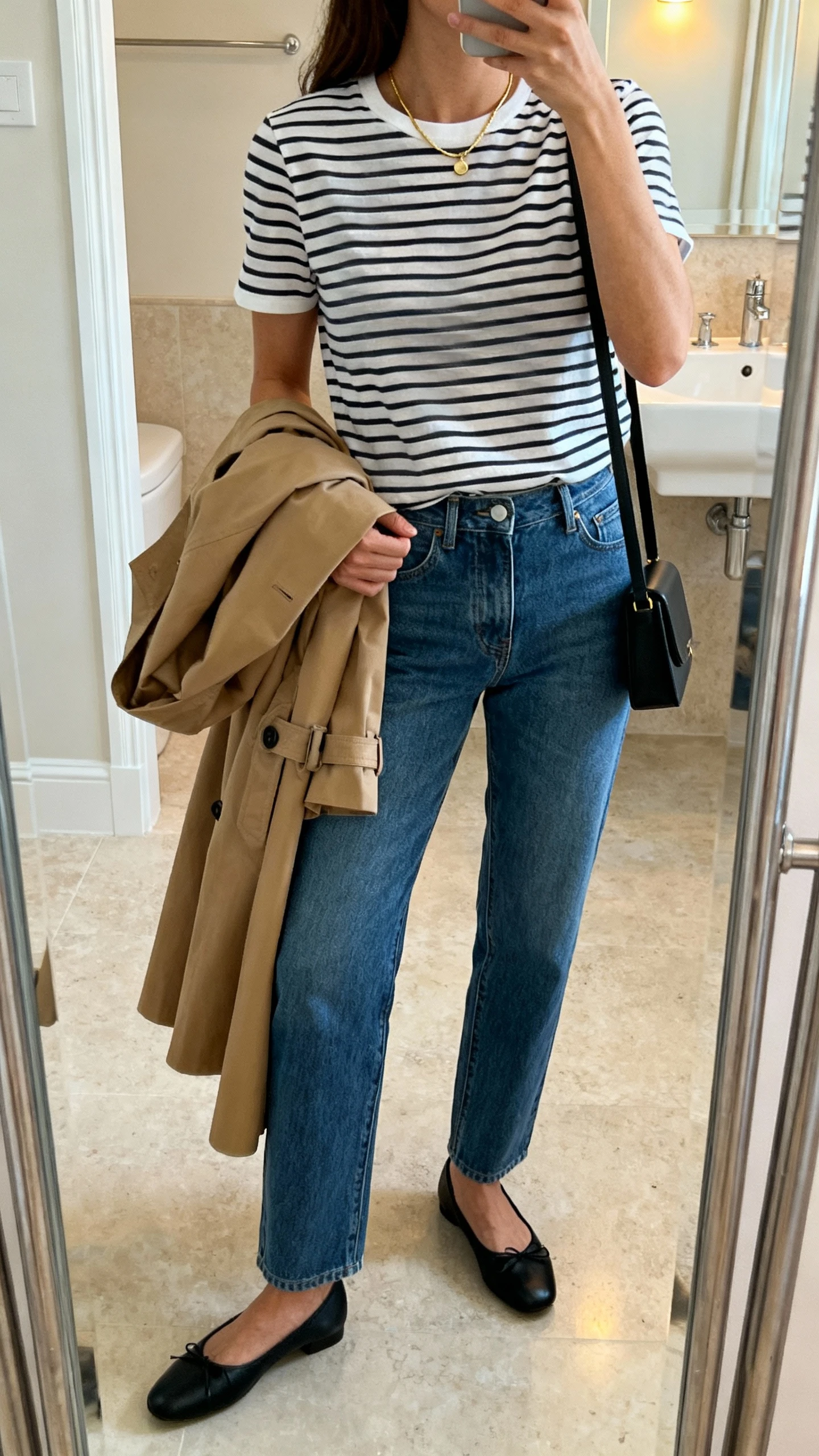 iPhone mirror selfie side angle of a woman wearing a classic striped Breton tee tucked into straight-leg blue jeans, black ballet flats, tan trench coat draped over arm, slim black shoulder bag, delicate gold necklace, face not visible, bathroom full-length mirror with soft indoor lighting, iPhone photo quality.
