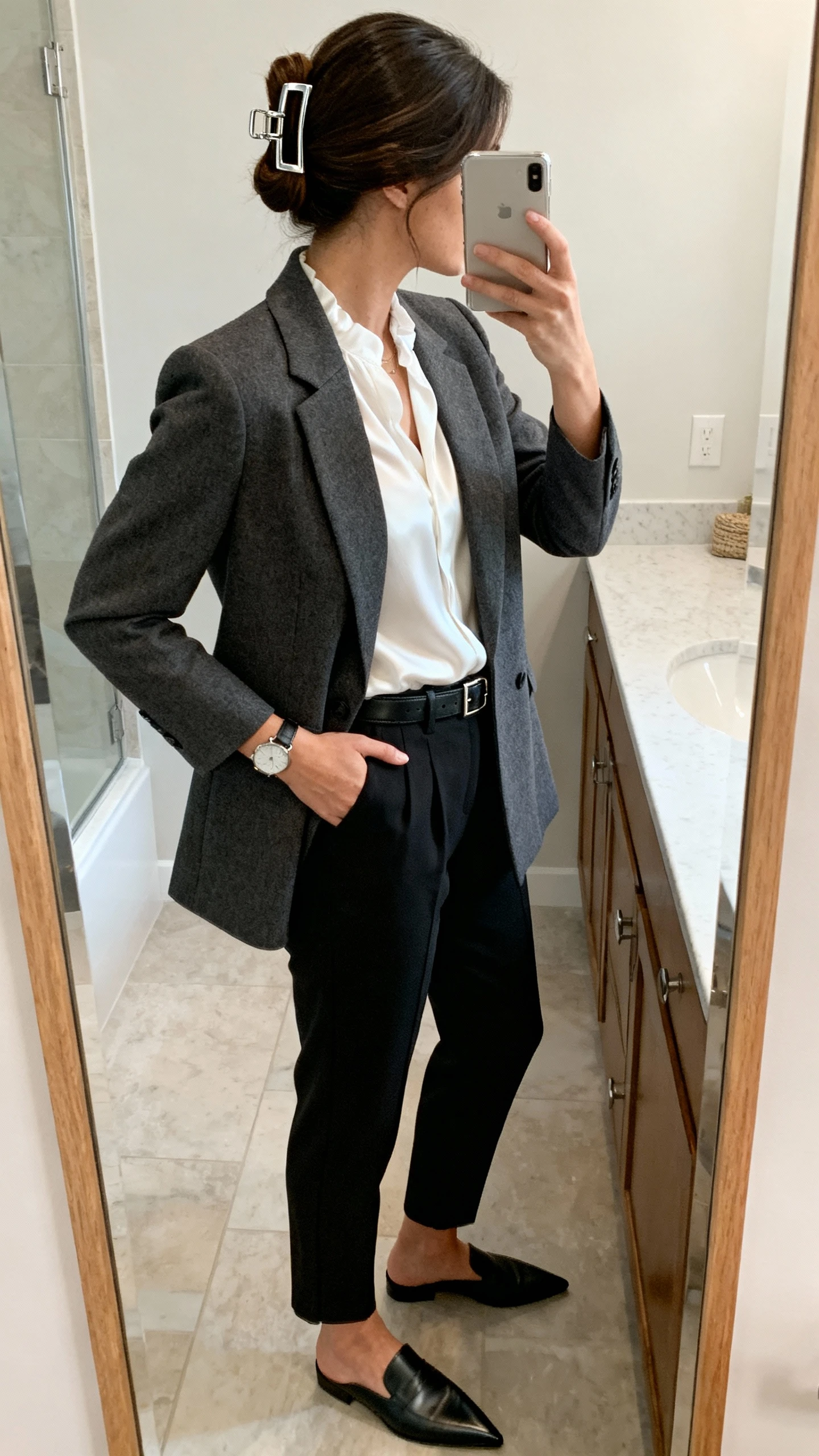 iPhone side-angle bathroom mirror selfie of a woman wearing a tailored charcoal blazer, white silk blouse, tapered black trousers, pointed-toe loafers, slim leather belt, minimalist watch, and hair swept up with a sleek metal claw clip; face not visible, natural indoor lighting, iPhone photo quality.