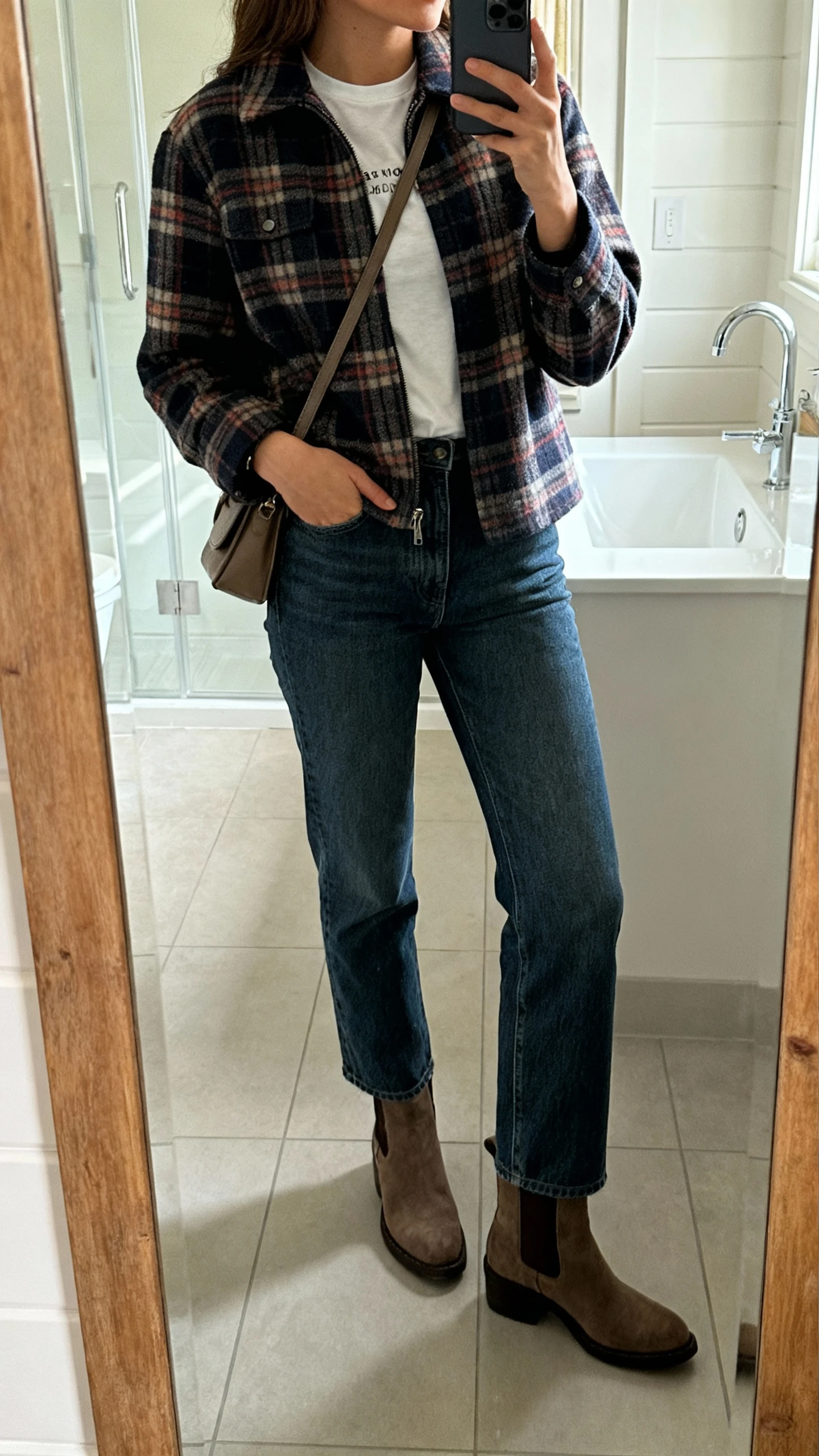 iPhone side-angle bathroom mirror selfie of a woman wearing a plaid shacket over a basic tee, straight-leg jeans, ankle boots, and a crossbody bag, face not visible, morning indoor light, casual iPhone photo.