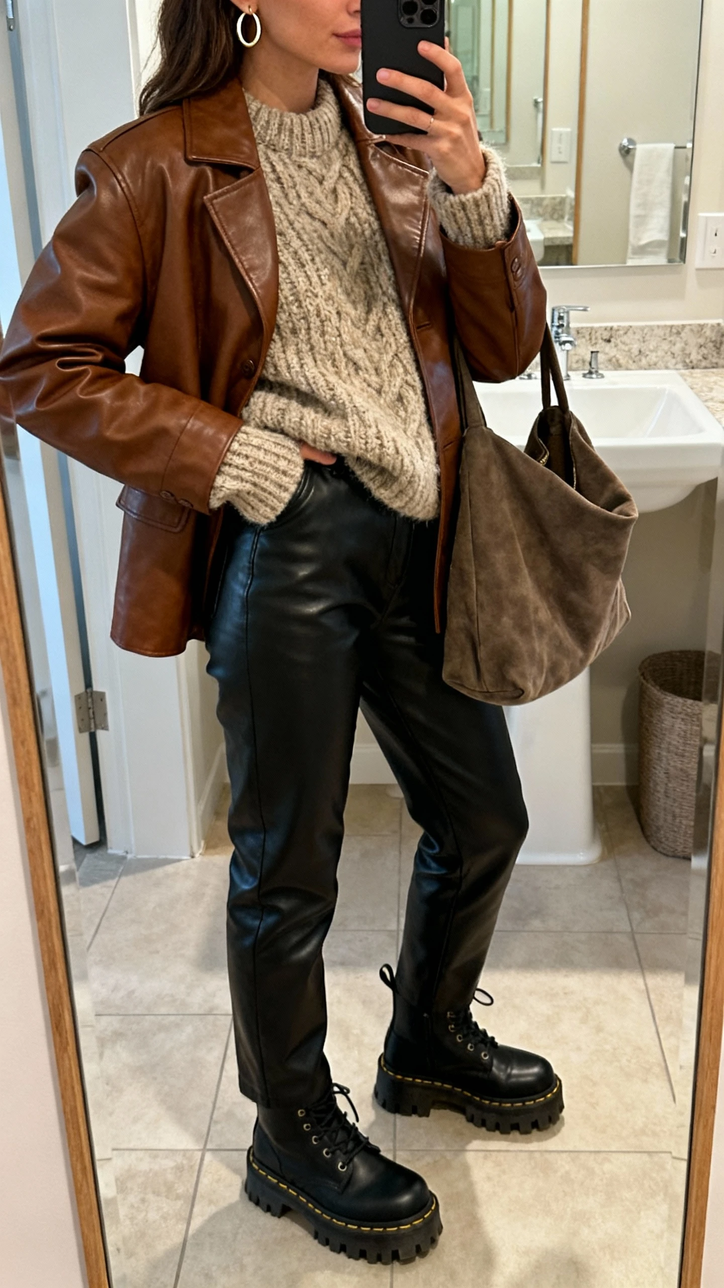 iPhone side-angle mirror selfie of a woman in a brown leather jacket with a chunky cozy knit sweater, black leather pants, lug-sole boots, hoop earrings, slouchy tote, face not visible, bathroom mirror, soft indoor lighting, iPhone photo quality