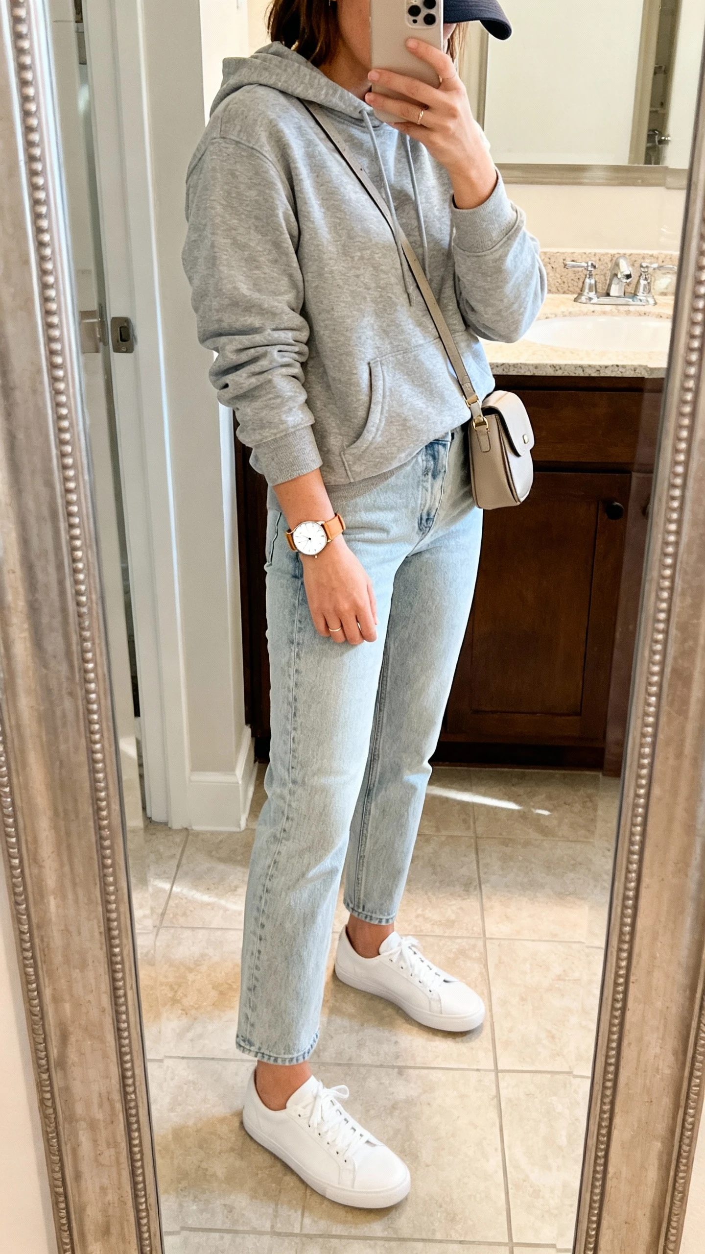 iPhone side-angle mirror selfie of a woman in a clean weekend uniform: grey hoodie, light-wash straight jeans, white low-top sneakers, minimalist tan leather watch with white dial, small crossbody bag, baseball cap, face not visible, bathroom mirror, soft morning light, iPhone photo quality.