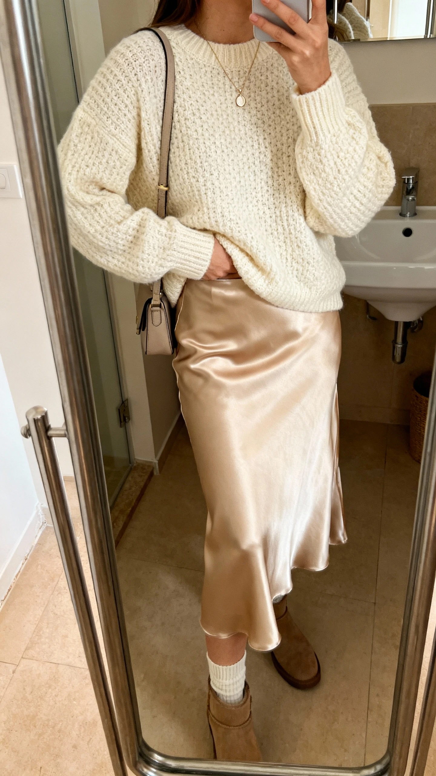 iPhone side-angle mirror selfie of a woman in a cozy cream chunky sweater half-tucked into a champagne satin midi skirt, ankle sock boots, dainty pendant necklace, crossbody bag, face not visible, bathroom full-length mirror, warm indoor lighting, iPhone photo quality.