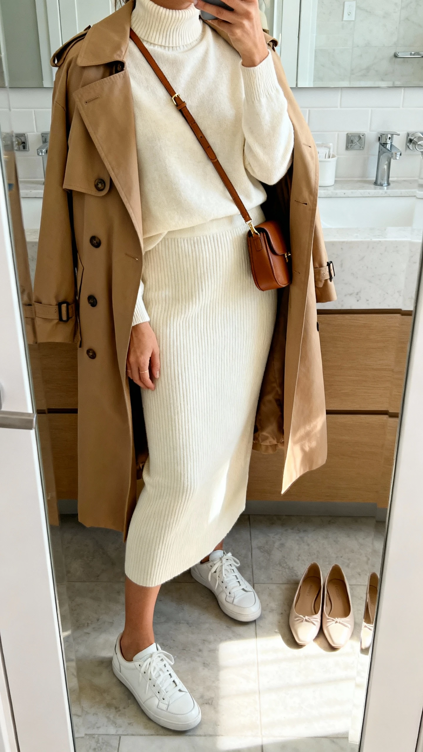 iPhone side-angle mirror selfie of a woman in a cream lightweight mock-neck knit top with matching cream knit midi skirt, camel trench coat draped over shoulders, white leather sneakers next to almond-toe ballet flats on the floor, minimalist leather crossbody; face not visible, bathroom mirror, soft morning light, iPhone photo quality.