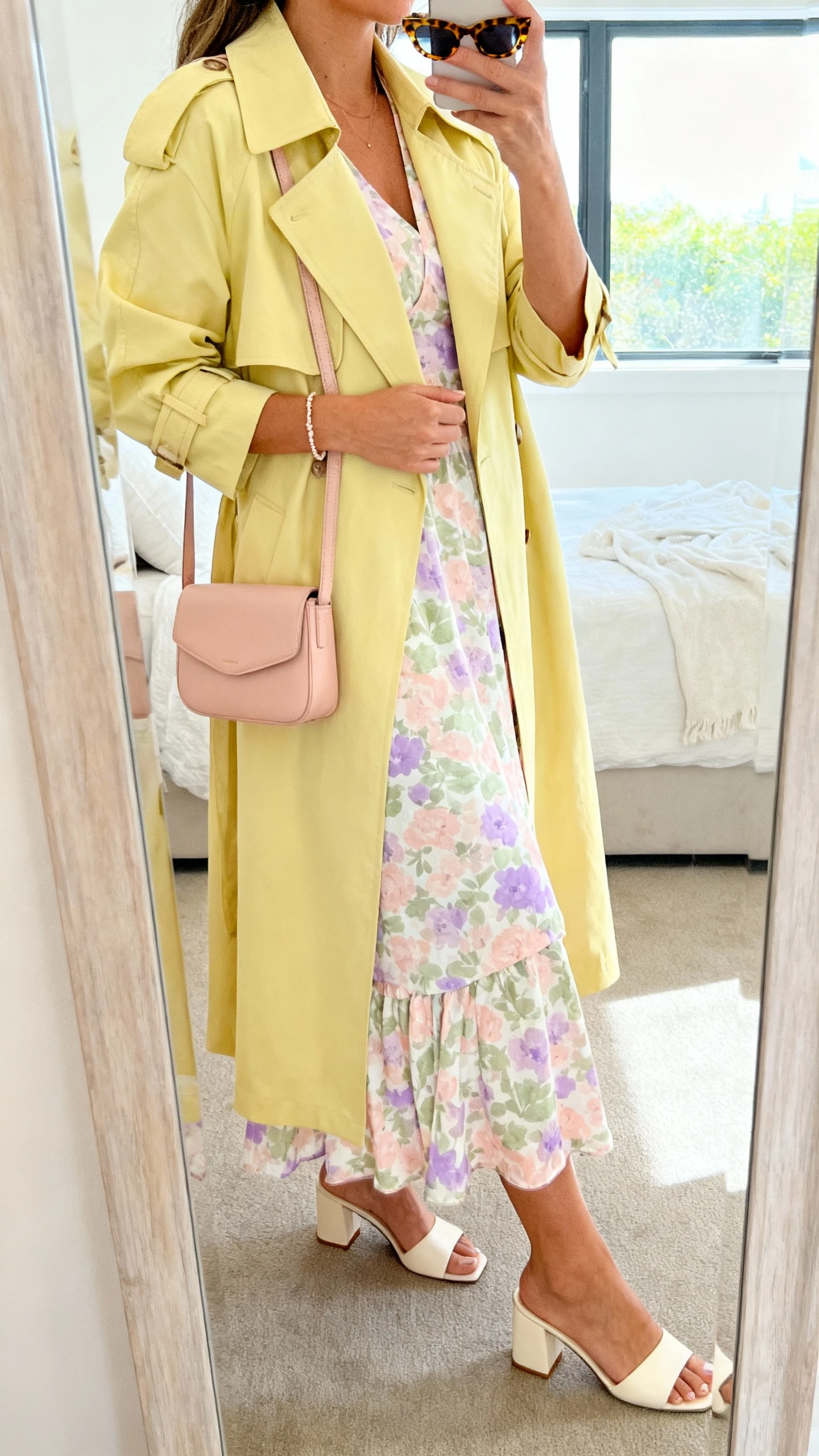 iPhone side-angle mirror selfie of a woman in a lightweight butter-yellow trench coat over a pastel floral midi dress (blush, sage, lilac accents), block-heel sandals in cream, a soft blush crossbody bag, tortoiseshell sunglasses, and a delicate bracelet, face not visible, bedroom mirror, bright window light, iPhone photo quality.