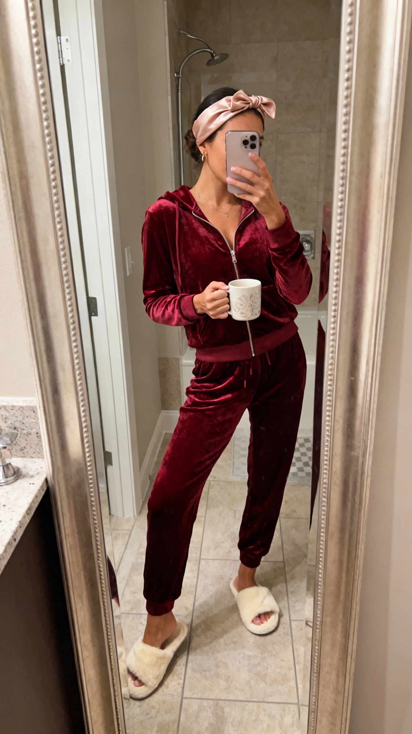 iPhone side-angle mirror selfie of a woman in a luxe velour lounge set: deep red velour zip-up hoodie and matching joggers, plush cream slippers, satin headband, holding a mug; face not visible, bathroom full-length mirror, warm indoor lighting, iPhone photo quality.