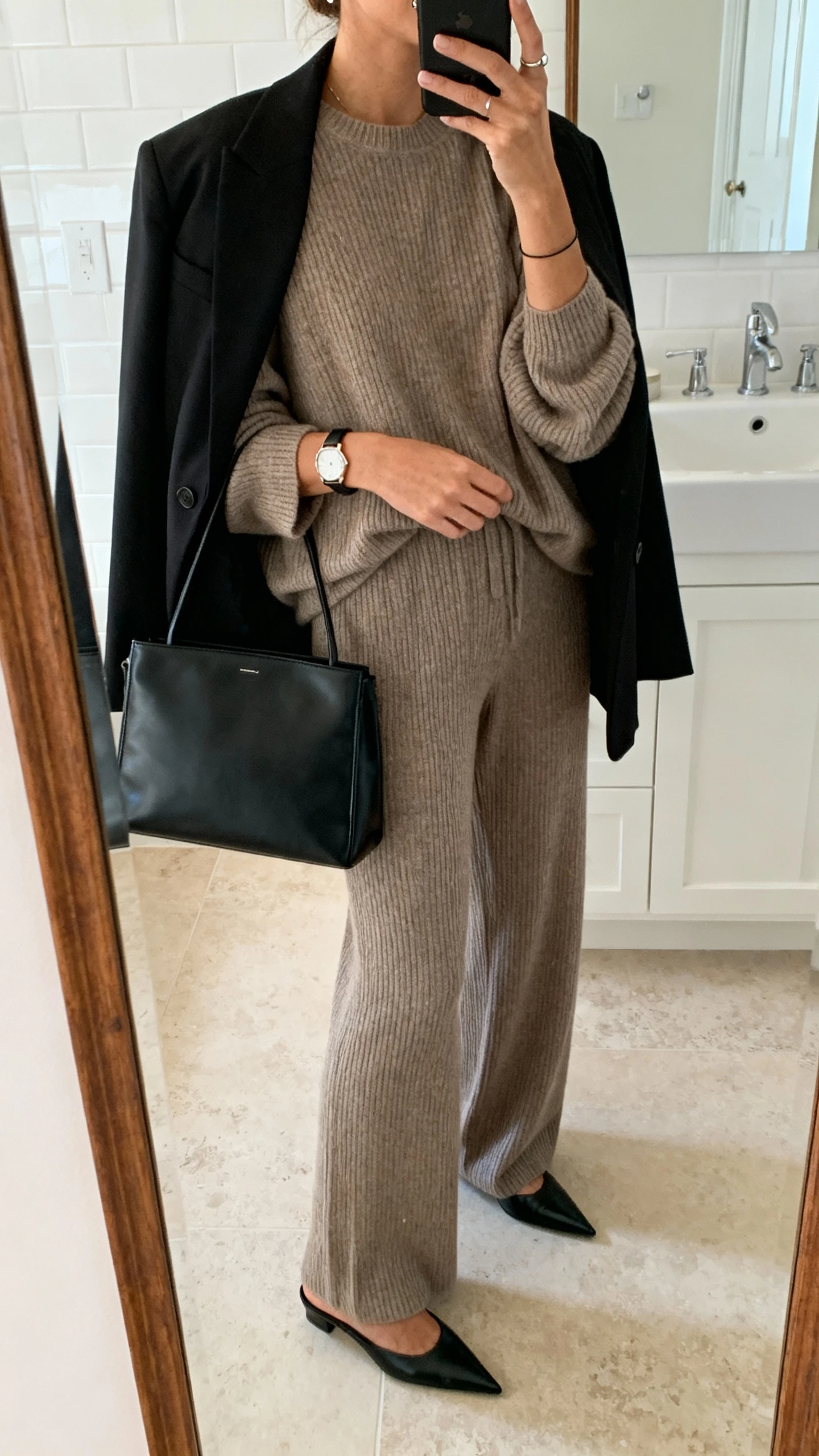 iPhone side-angle mirror selfie of a woman in a matching taupe ribbed knit set (slouchy crewneck and wide-leg knit pants), sleek black blazer draped over shoulders, minimalist black leather tote, pointed-toe black mules, thin watch and simple studs, face not visible, bathroom full-length mirror, soft morning light, iPhone photo quality.