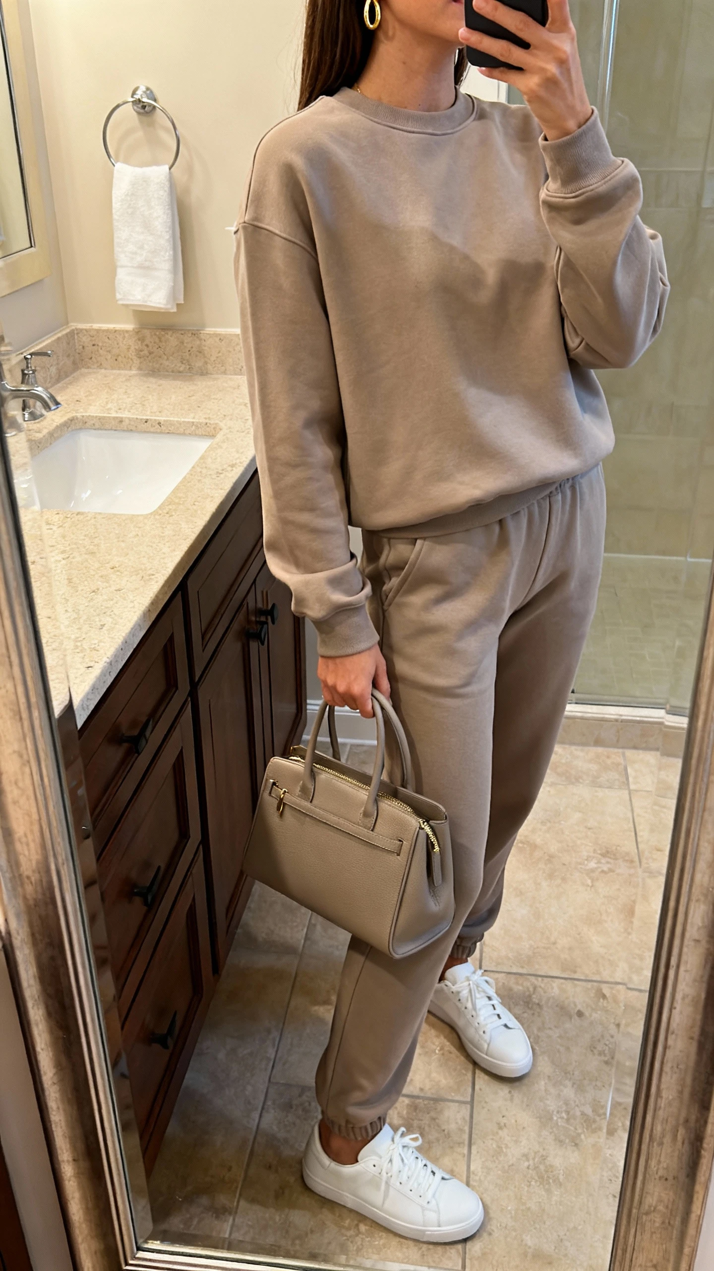 iPhone side-angle mirror selfie of a woman in a matching elevated sweat set (tailored sweatshirt and joggers), clean white sneakers, minimal gold hoops, and a structured tote; face not visible, bathroom mirror, warm indoor lighting, iPhone photo quality.