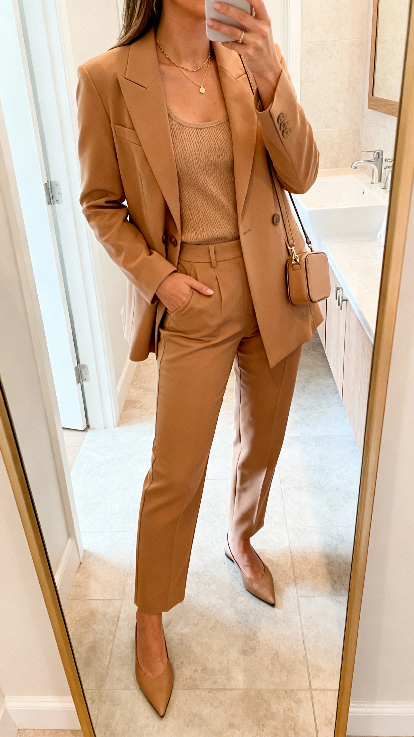 iPhone side-angle mirror selfie of a woman in a monochrome camel set: matching tailored blazer and straight-leg trousers with a tonal knit tank, delicate necklace, pointed flats, and a compact crossbody, face not visible, bathroom mirror, bright indoor lighting, iPhone photo quality.