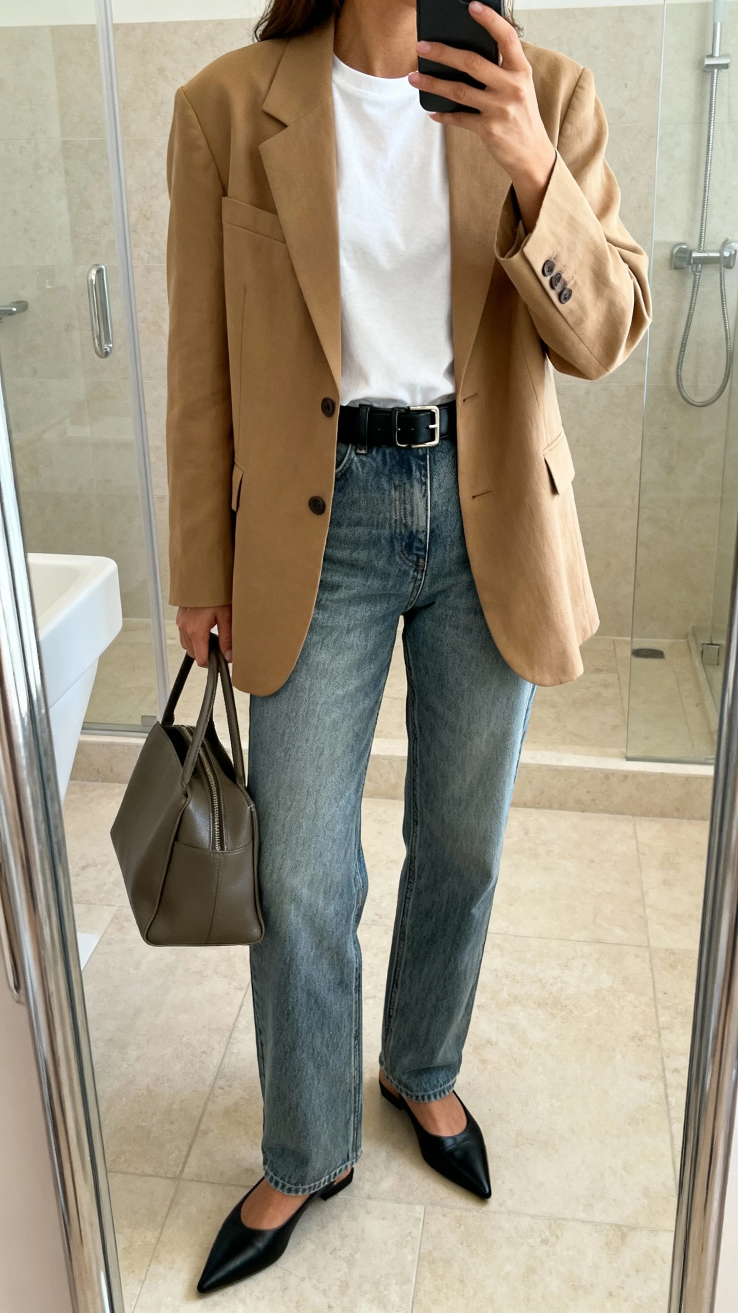 iPhone side-angle mirror selfie of a woman in a relaxed camel blazer over a white tee, straight-leg medium-wash jeans, black leather belt, pointed black flats, and a structured tote, face not visible, cropped at shoulders, bathroom full-length mirror, soft indoor lighting, iPhone photo quality