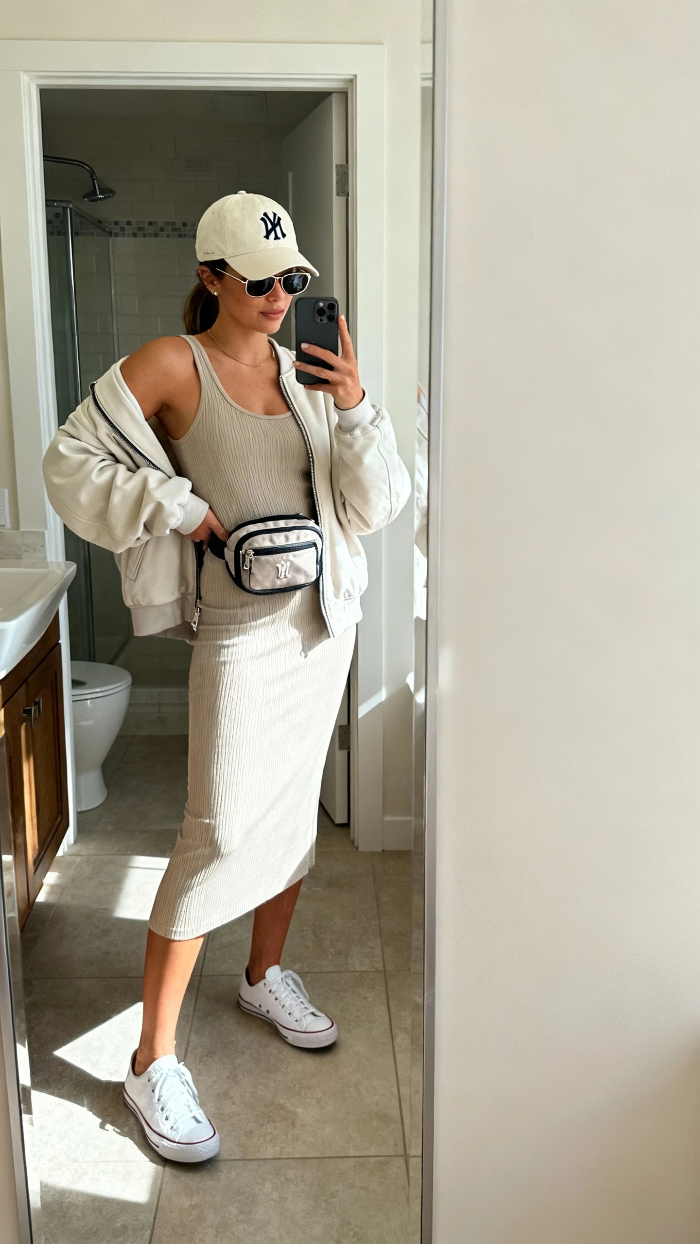 iPhone side-angle mirror selfie of a woman in a ribbed tank dress (knee-length or midi), white low-top sneakers (leather or canvas), a neutral or team-logo baseball cap, a lightweight zip-up hoodie or oversized bomber jacket, a mini backpack or belt bag, and sleek-frame sunglasses, face not visible, bathroom mirror, morning light, iPhone photo quality.