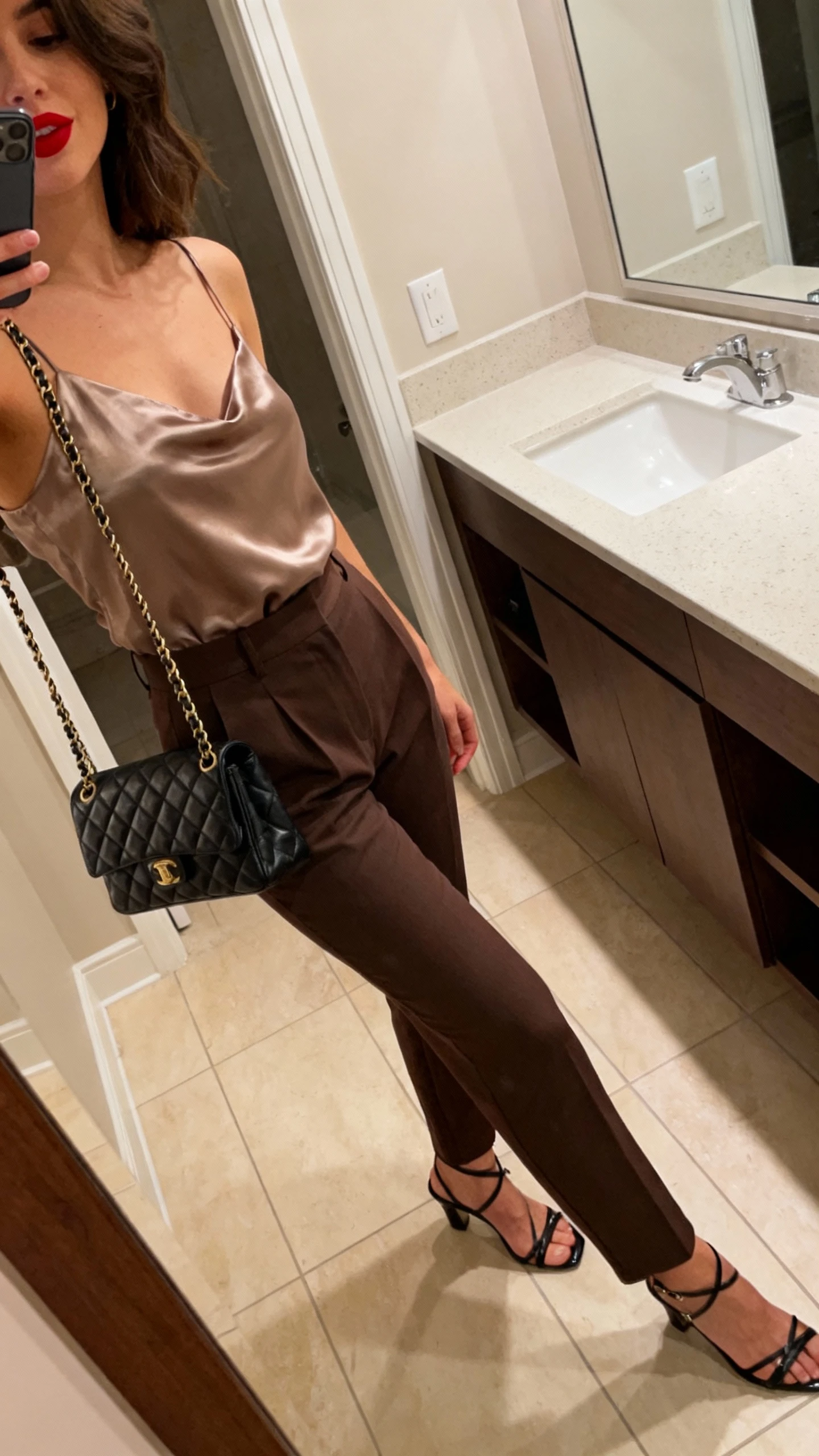 iPhone side-angle mirror selfie of a woman in a satin camisole, high-waist trousers, strappy heels, red lip glimpsed off-frame, and a quilted chain shoulder bag, face not visible, bathroom mirror with overhead warm lighting, iPhone photo quality.