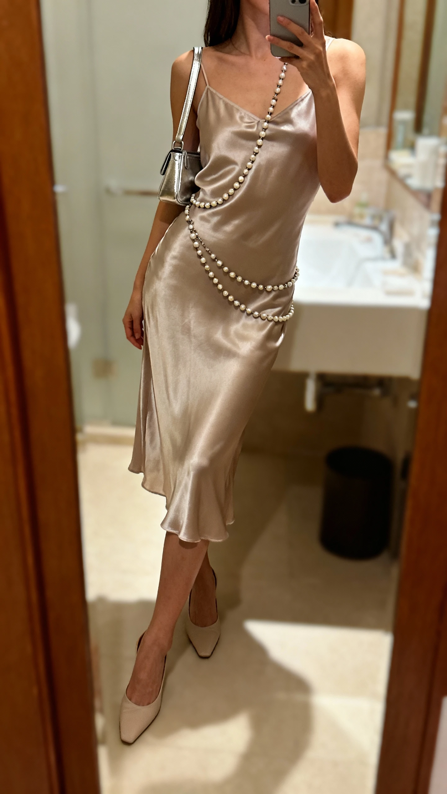 iPhone side-angle mirror selfie of a woman in a satin slip dress with a beaded body chain draped over it, minimalist heels and a small metallic shoulder bag, face not visible, bathroom mirror, warm indoor lighting, iPhone photo quality.