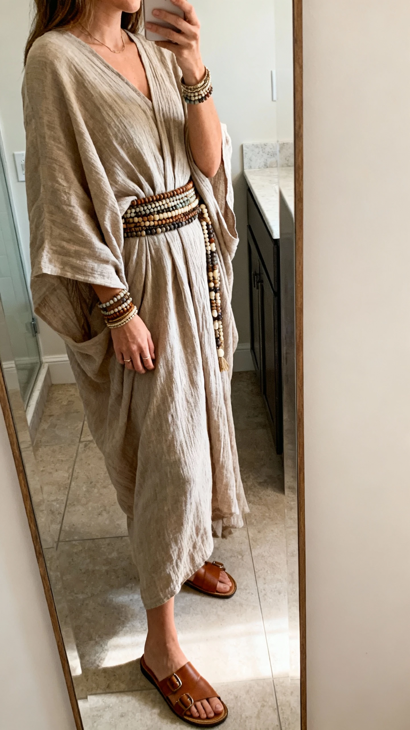 iPhone side-angle mirror selfie of a woman in a slouchy kaftan cinched with stacked beaded belts, flat leather sandals and bangles, face not visible, bathroom full-length mirror, soft indoor morning light, iPhone photo quality.