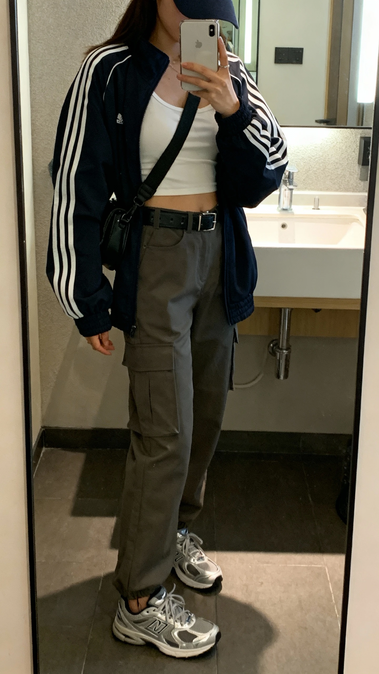 iPhone side-angle mirror selfie of a woman in a sporty-city mix: oversized track jacket, fitted tank, high-waisted cargo pants, sleek sneakers, crossbody belt bag, and a cap, face not visible, bathroom mirror with indoor lighting, iPhone photo quality.