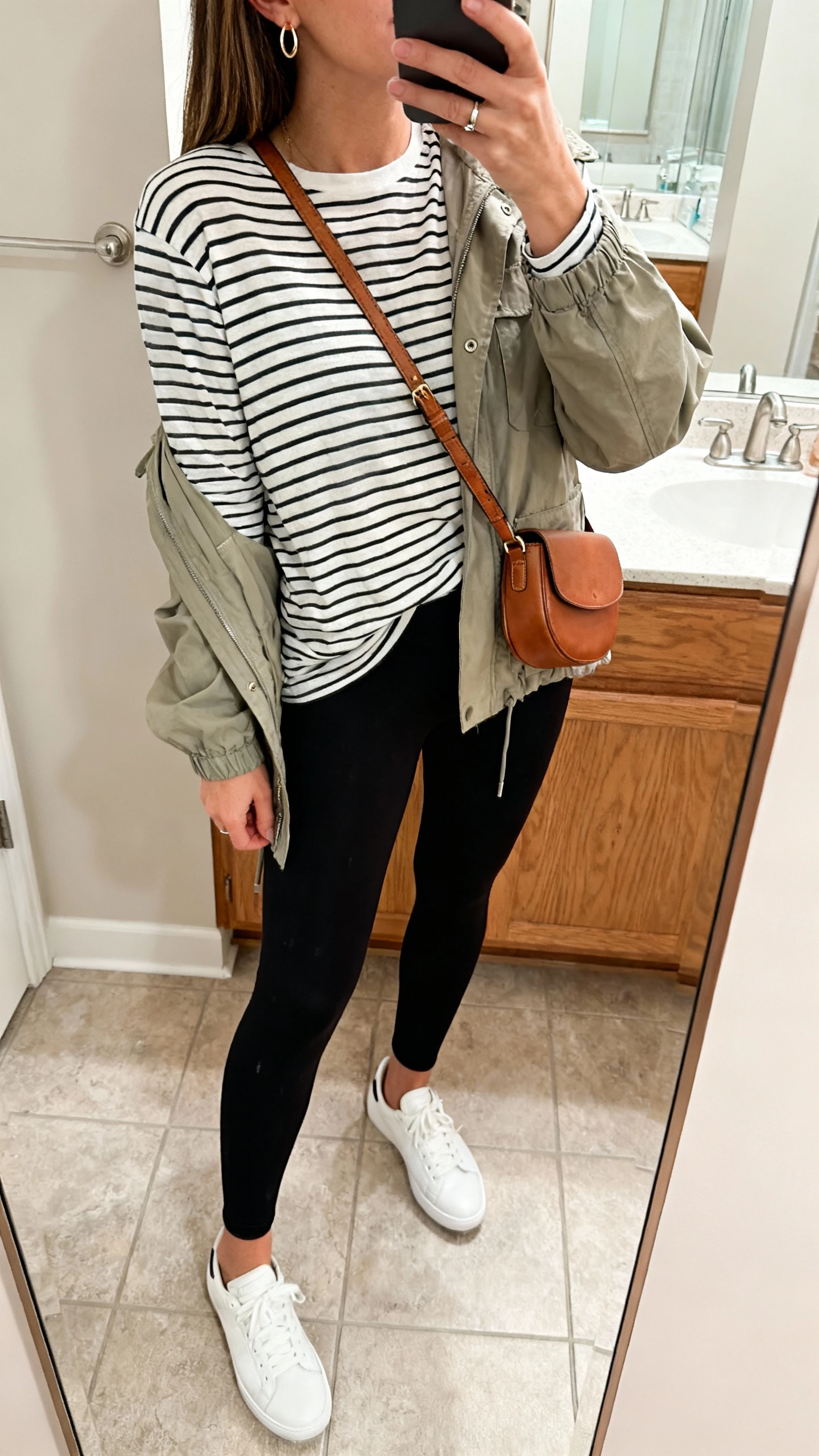 iPhone side-angle mirror selfie of a woman in a striped long-sleeve tee, black ankle-length leggings, lightweight utility jacket, white sneakers, hands-free leather crossbody worn across the chest, small hoop earrings and simple ring; face not visible, cropped at shoulders, bathroom mirror, natural indoor lighting, iPhone photo quality.