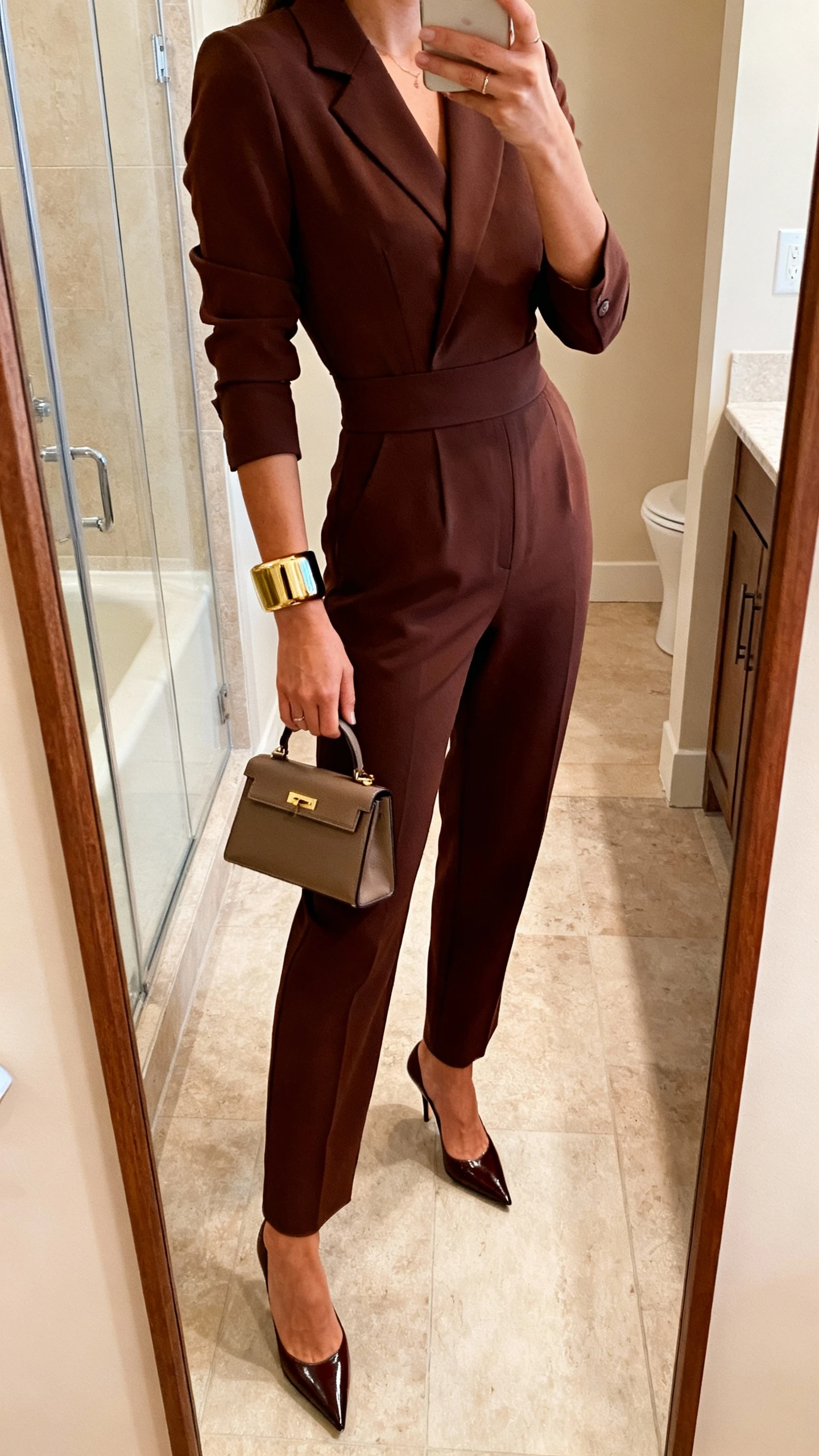 iPhone side-angle mirror selfie of a woman in a tailored power jumpsuit with a defined waist, pointed-toe heels, structured mini handbag, and bold cuff bracelet, face not visible, bathroom full-length mirror, warm indoor lighting, iPhone photo quality.