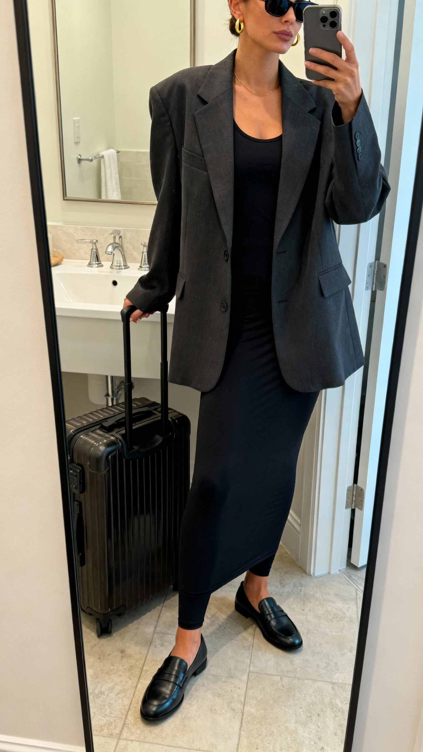 iPhone side-angle mirror selfie of a woman in an oversized blazer over a longline black leggings set (tank or tee), sleek slip-on loafers, structured carry-on tote, slim sunglasses, and dainty gold hoops; face not visible, cropped at shoulders, bathroom mirror, indoor soft lighting, iPhone photo quality.