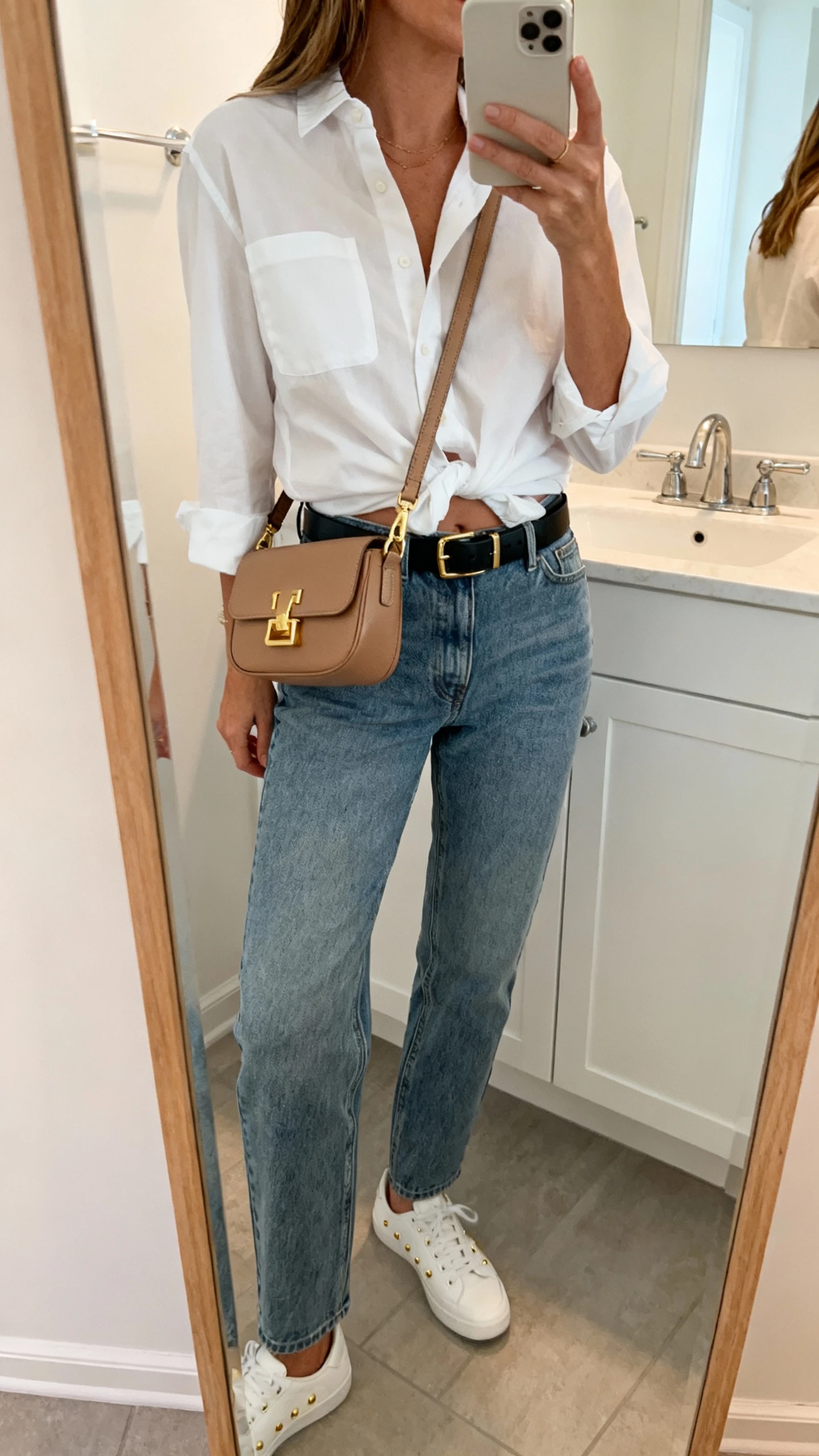 iPhone side-angle mirror selfie of a woman in mid-wash straight-leg jeans, relaxed white button-down half-tucked, black leather belt, minimalist white sneakers, subtle gold studs, wearing a luxe tan crossbody with gold hardware across the torso, face not visible, bathroom mirror, soft morning light, iPhone photo quality.