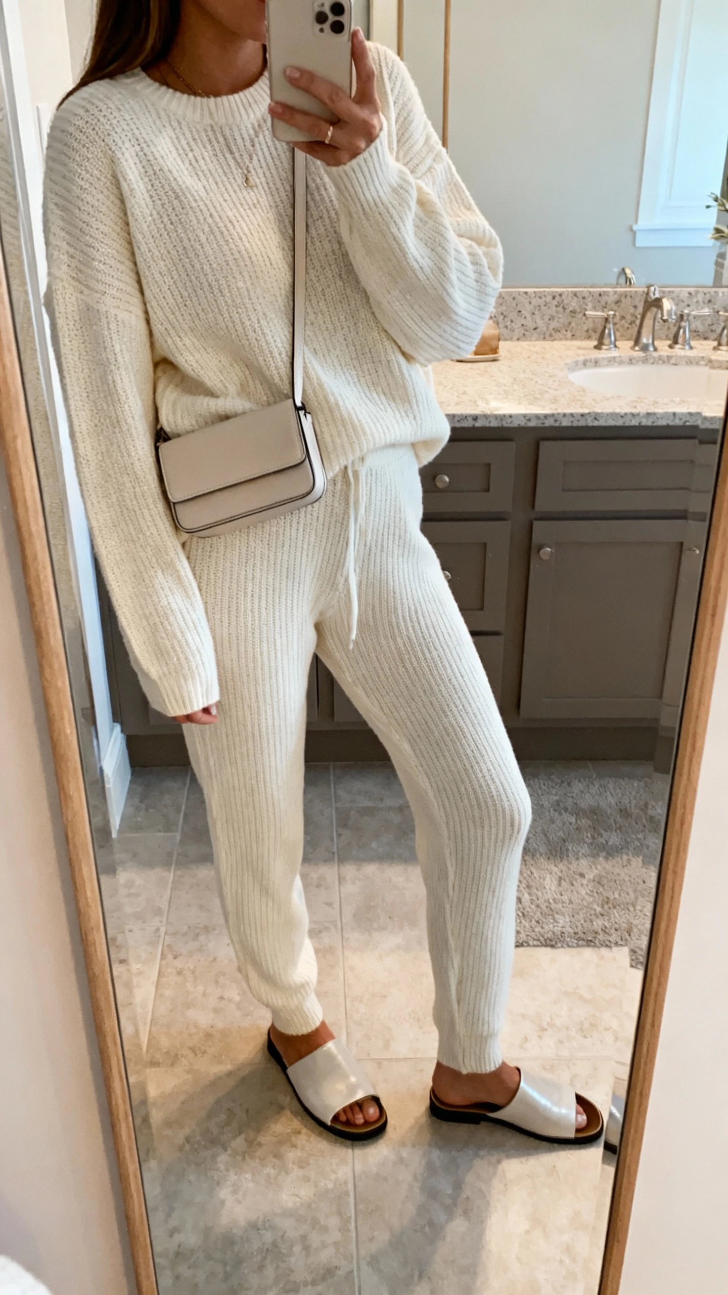 iPhone side-angle mirror selfie of a woman wearing a monochrome matching knit sweater and knit pants set with sleek slip-on mules and a compact crossbody, face not visible, cropped at shoulders, bathroom mirror, soft indoor lighting, iPhone photo quality.