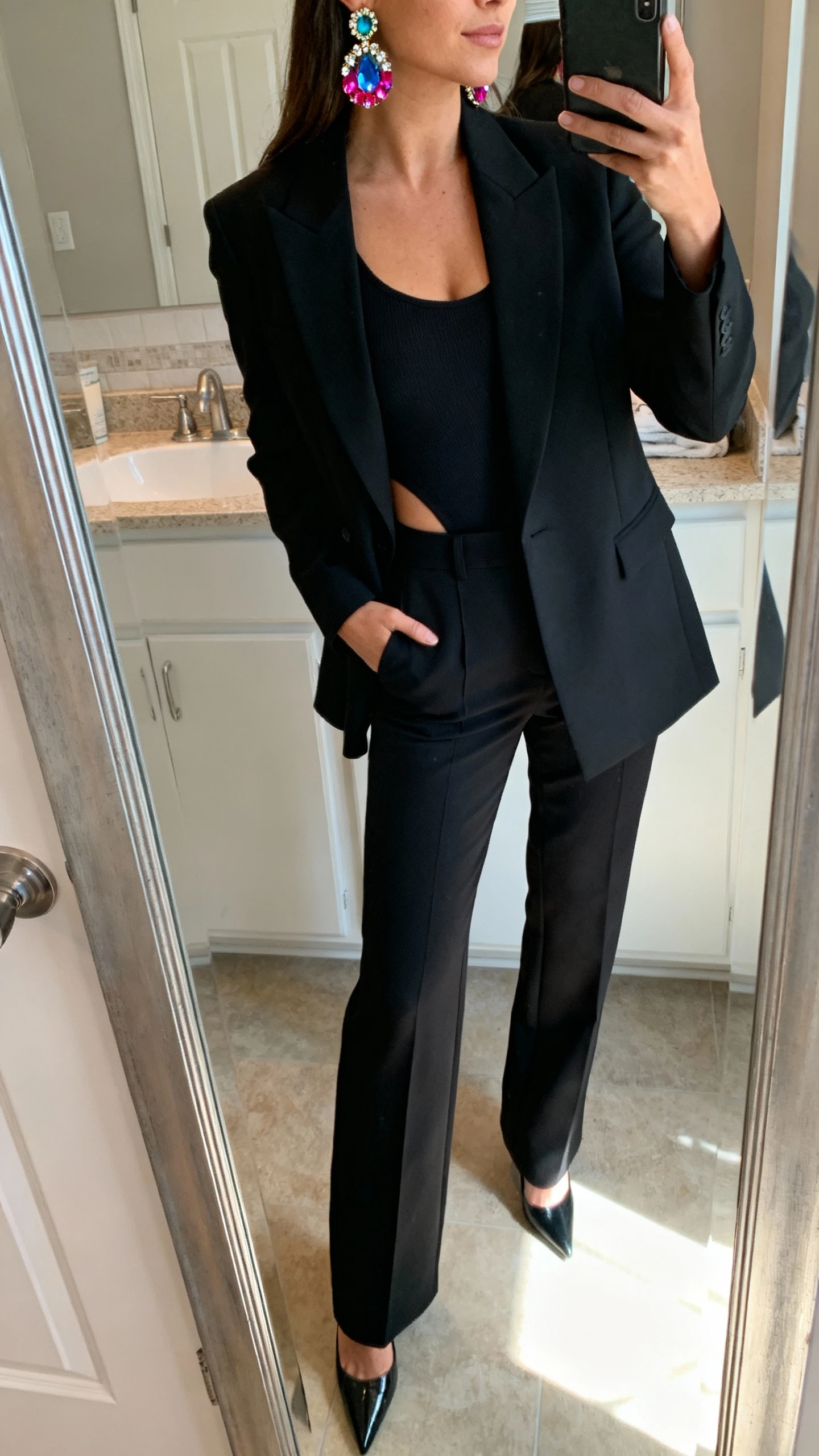 iPhone side-angle mirror selfie of a woman wearing a sharp black tailored blazer over a black bodysuit, high-rise black trousers, pointed heels, and bold color-pop gemstone statement earrings, face not visible, bathroom mirror, indoor morning light, iPhone photo quality.