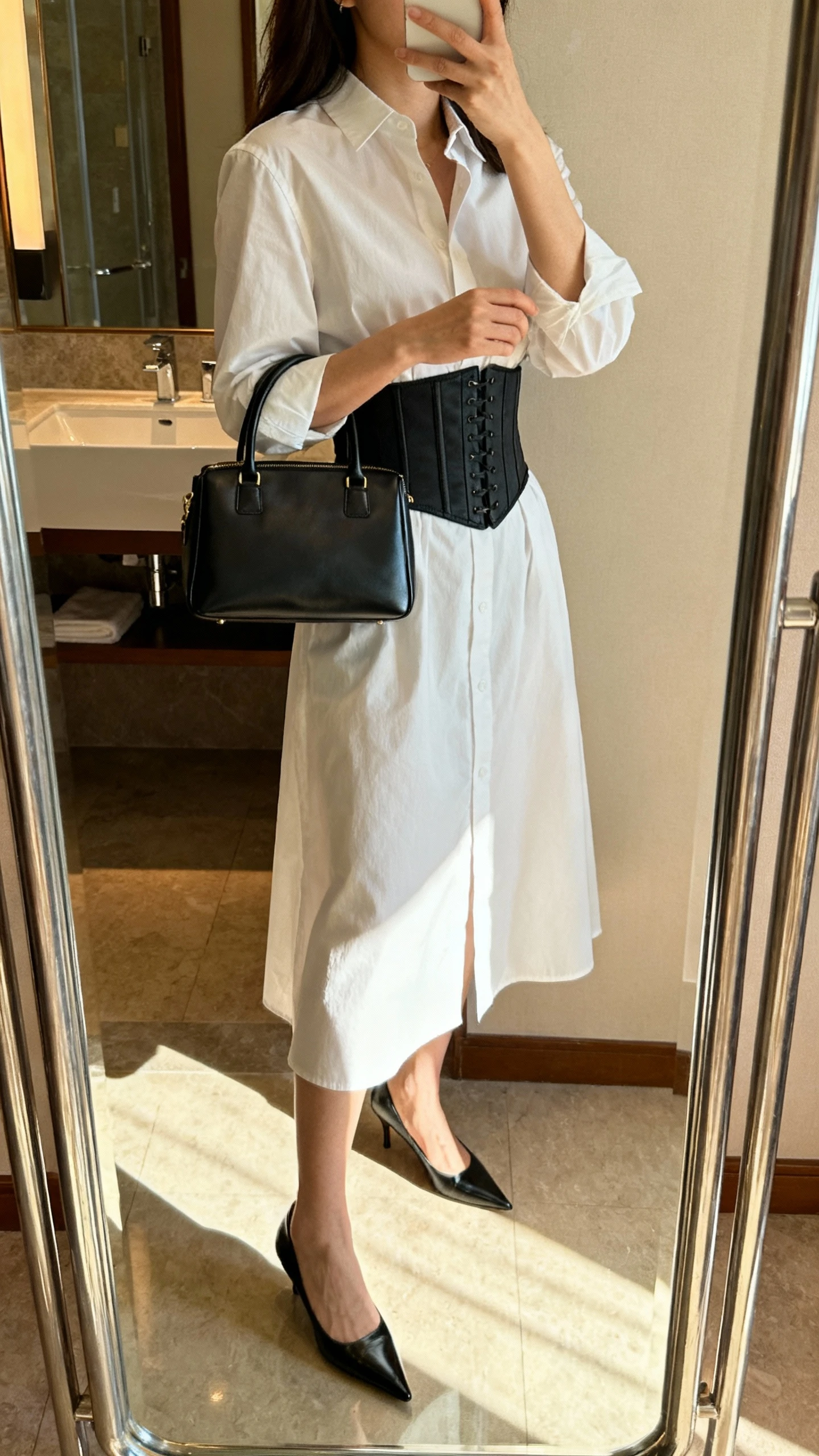 iPhone side-angle mirror selfie of a woman wearing a crisp shirt dress cinched with a corset belt, pointed-toe heels, sleek top-handle bag, face not visible, bathroom mirror with morning light, iPhone photo quality.