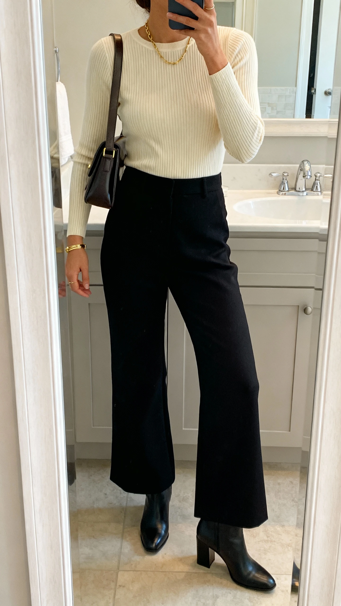 iPhone side-angle mirror selfie of a woman wearing a fitted ribbed sweater in cream with high-waisted wide-leg trousers in black, block-heel ankle boots, a structured shoulder bag, and simple gold jewelry; face not visible, bathroom full-length mirror, soft indoor lighting, iPhone photo quality.
