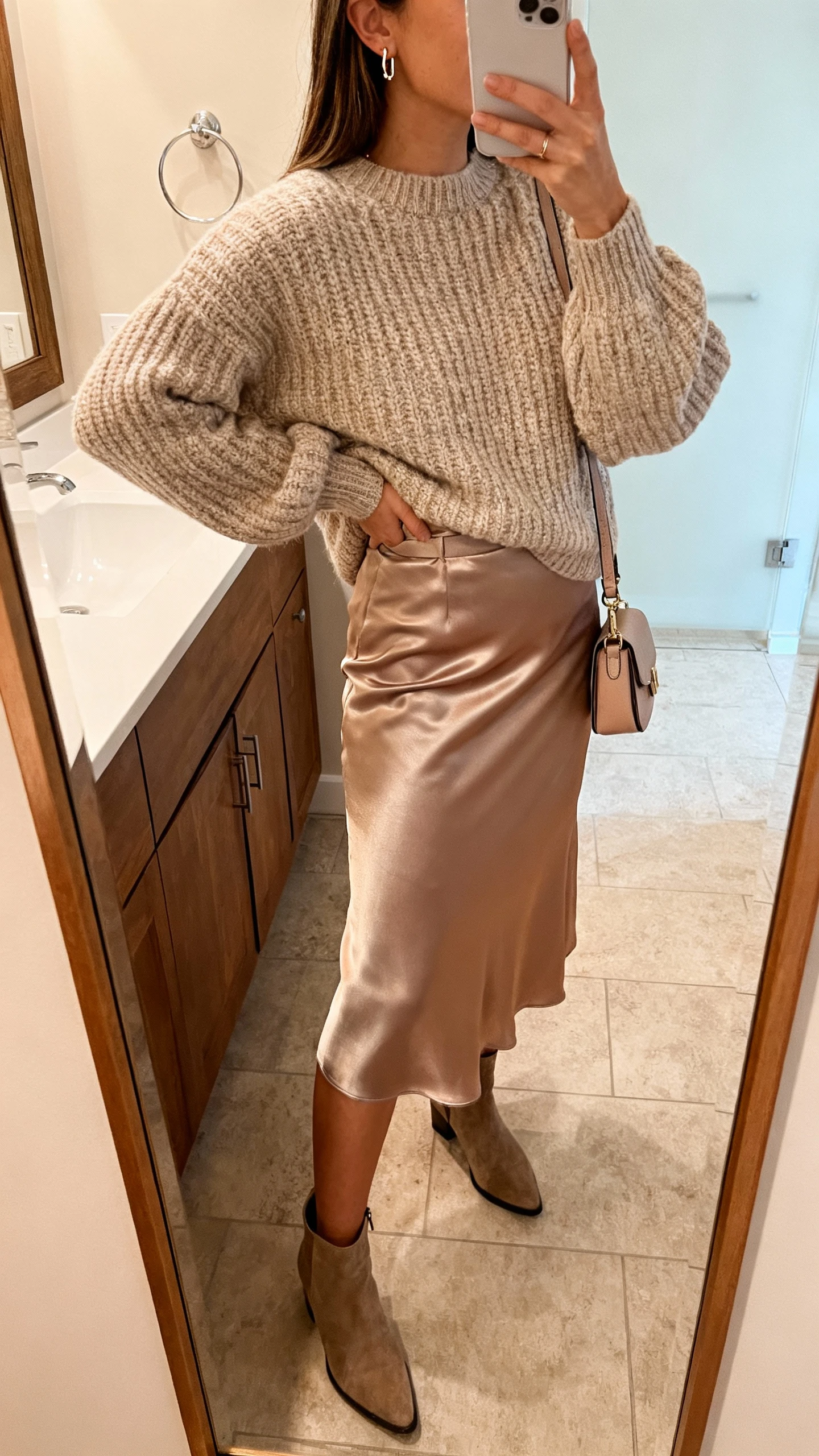 iPhone side-angle mirror selfie of a woman wearing a cozy chunky sweater tucked into a satin midi skirt with dainty earrings, ankle boots, and a mini shoulder bag, face not visible, bathroom mirror, warm indoor lighting, iPhone photo quality.