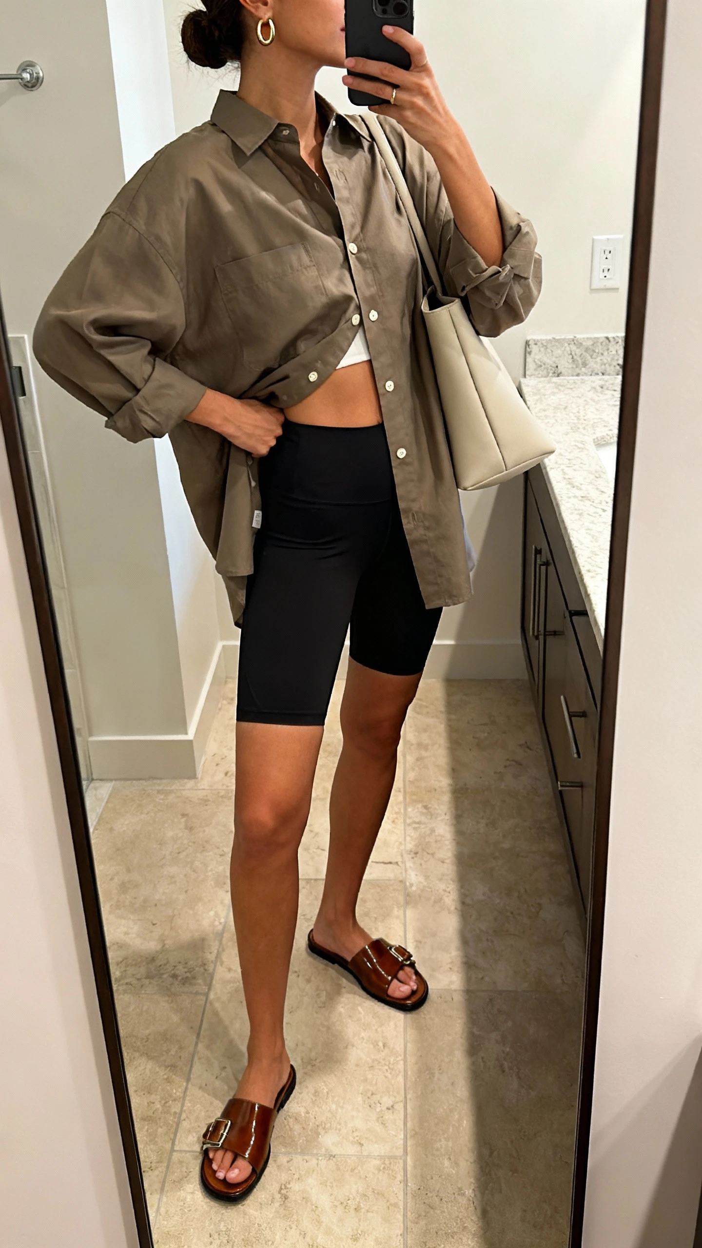 iPhone side-angle mirror selfie of a woman wearing oversized button-up shirt half-tucked into biker shorts, polished slide sandals, minimalist tote, dainty hoop earrings, face not visible, bathroom full-length mirror, soft indoor lighting, iPhone photo quality