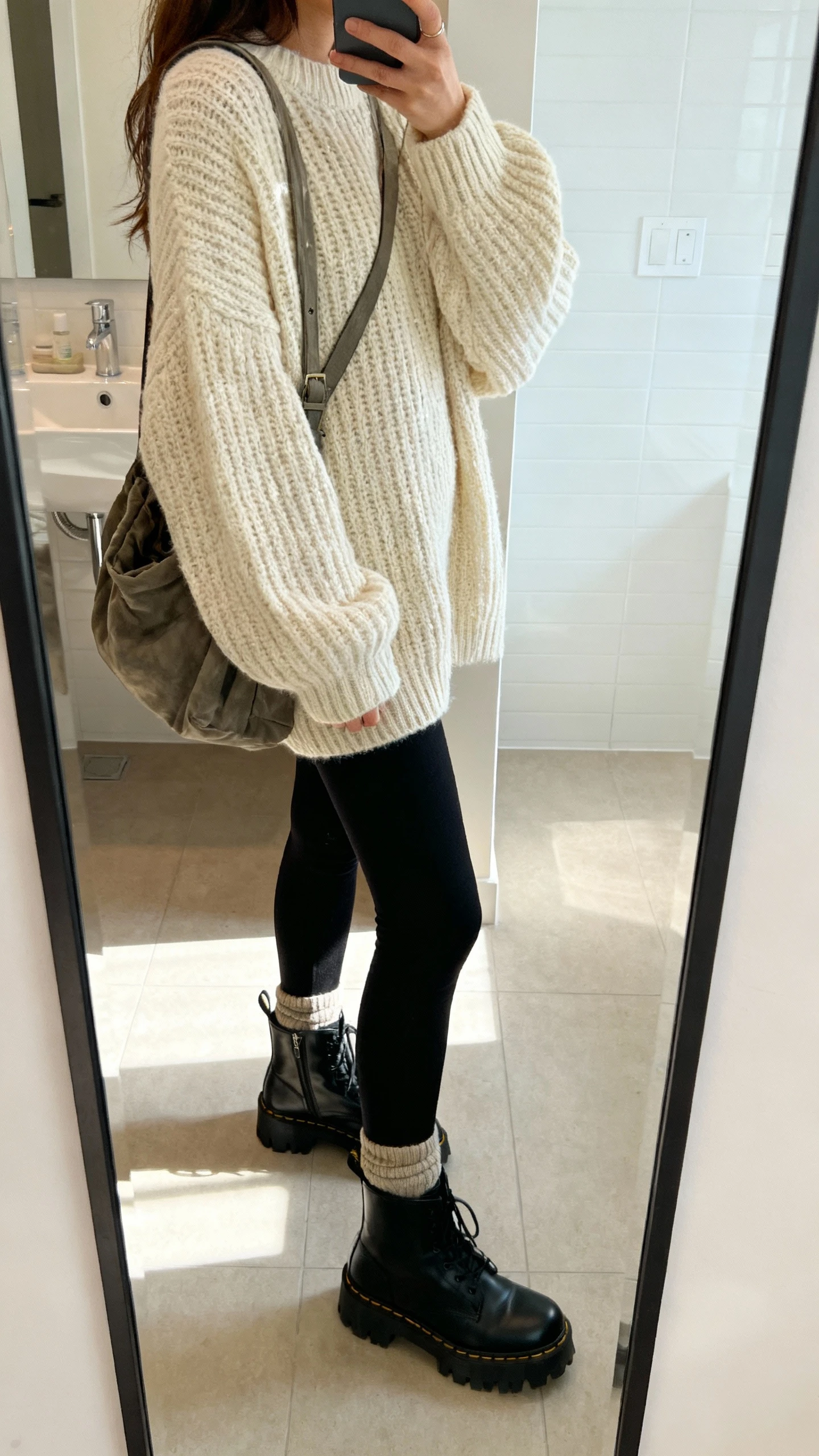 iPhone side-angle mirror selfie of a woman wearing black leggings, an oversized cream chunky knit sweater, black lug-sole ankle boots, thick socks peeking, and a slouchy crossbody, face not visible, bathroom mirror, morning indoor light, iPhone photo quality.