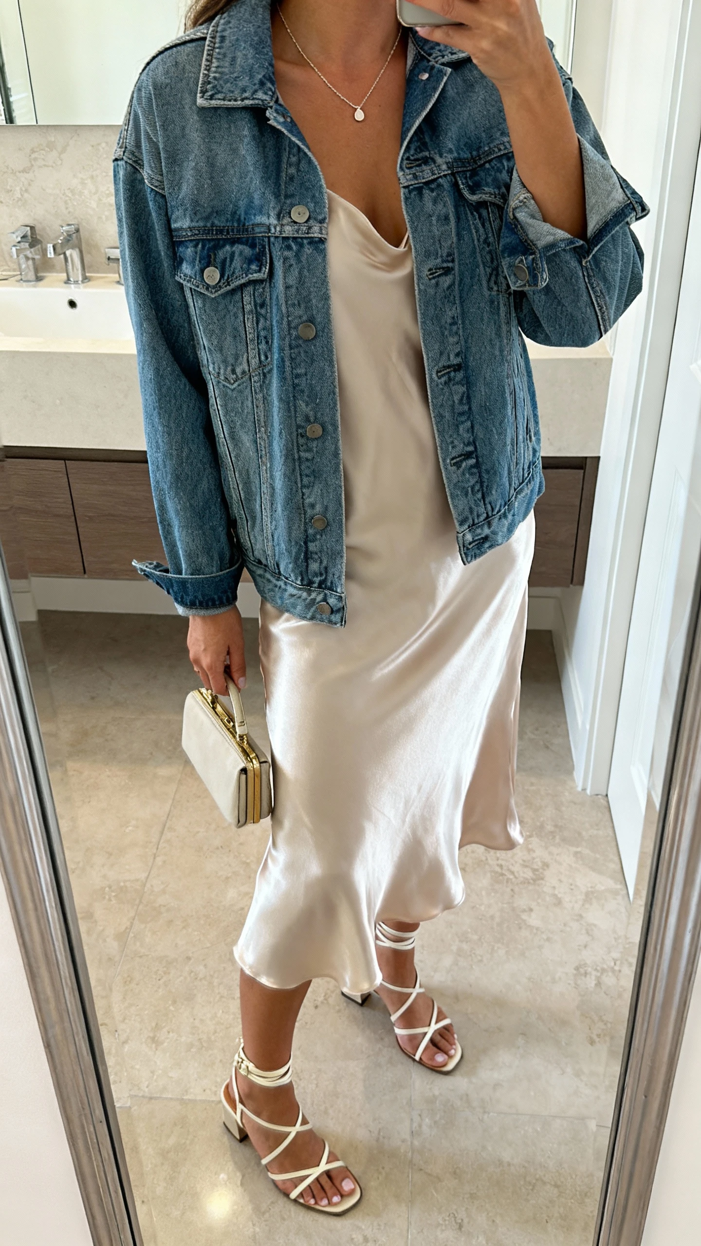 iPhone side-angle mirror selfie of a woman wearing a denim jacket layered over a silky slip dress with delicate necklace, small clutch, and strappy heeled sandals, face not visible, cropped at shoulders, bathroom mirror, soft indoor lighting, iPhone photo quality
