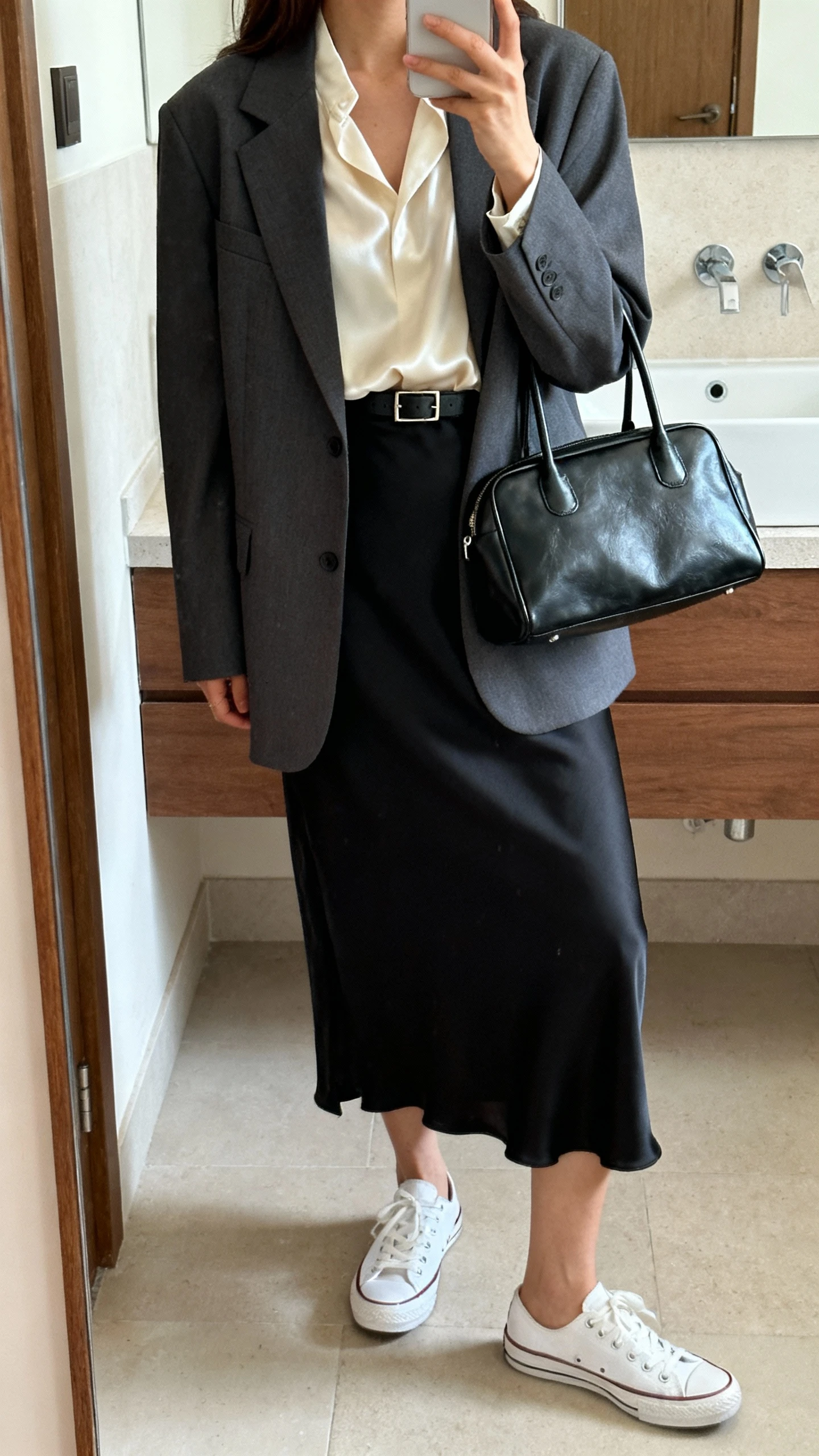 iPhone side-angle mirror selfie of a woman wearing white court-style sneakers, a charcoal gray blazer over a silk cream blouse, high-waisted black midi slip skirt, structured black leather tote, and a slim belt, face not visible, bathroom mirror with soft indoor lighting, iPhone photo quality.